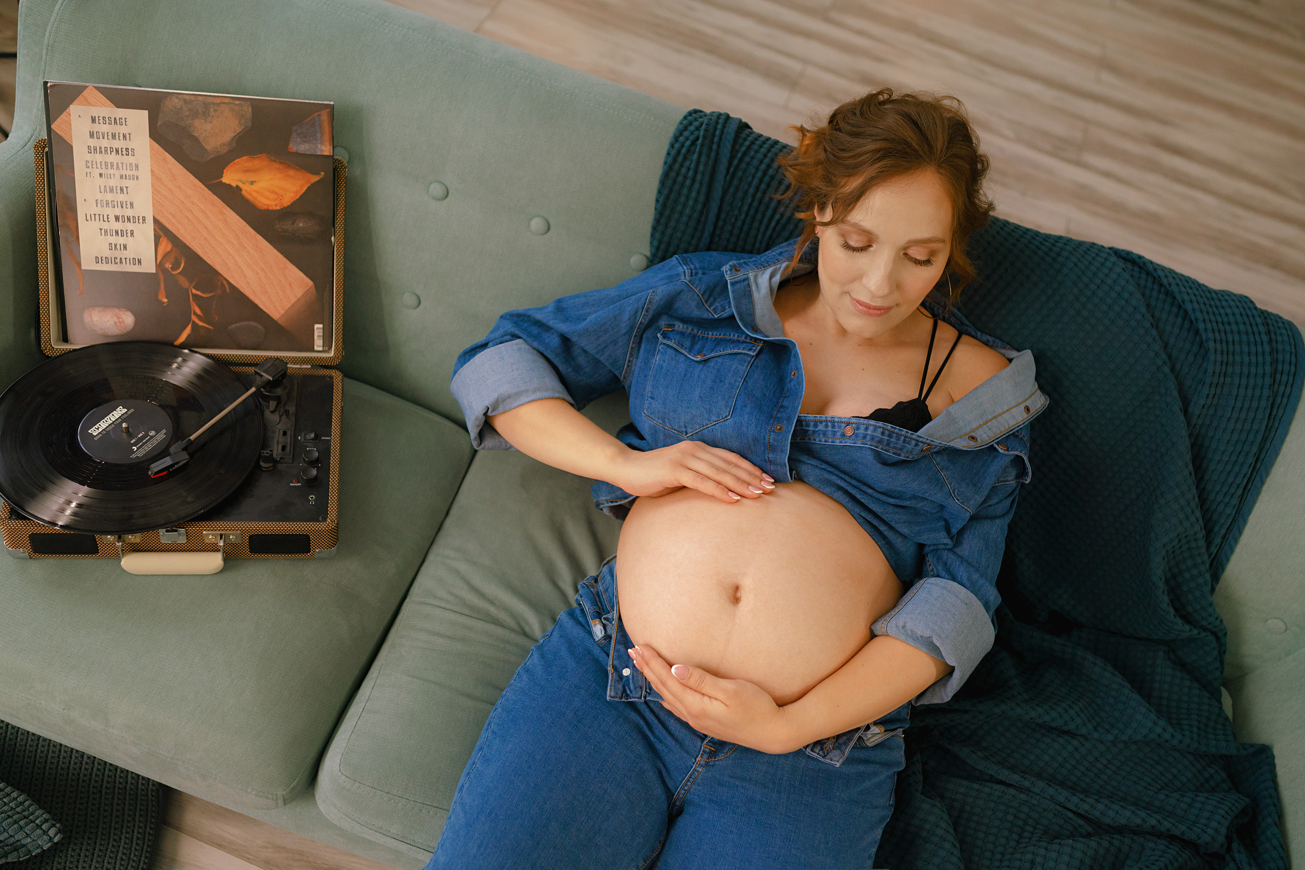 Беременная держится за живот и слушает грамафон сидя на диване