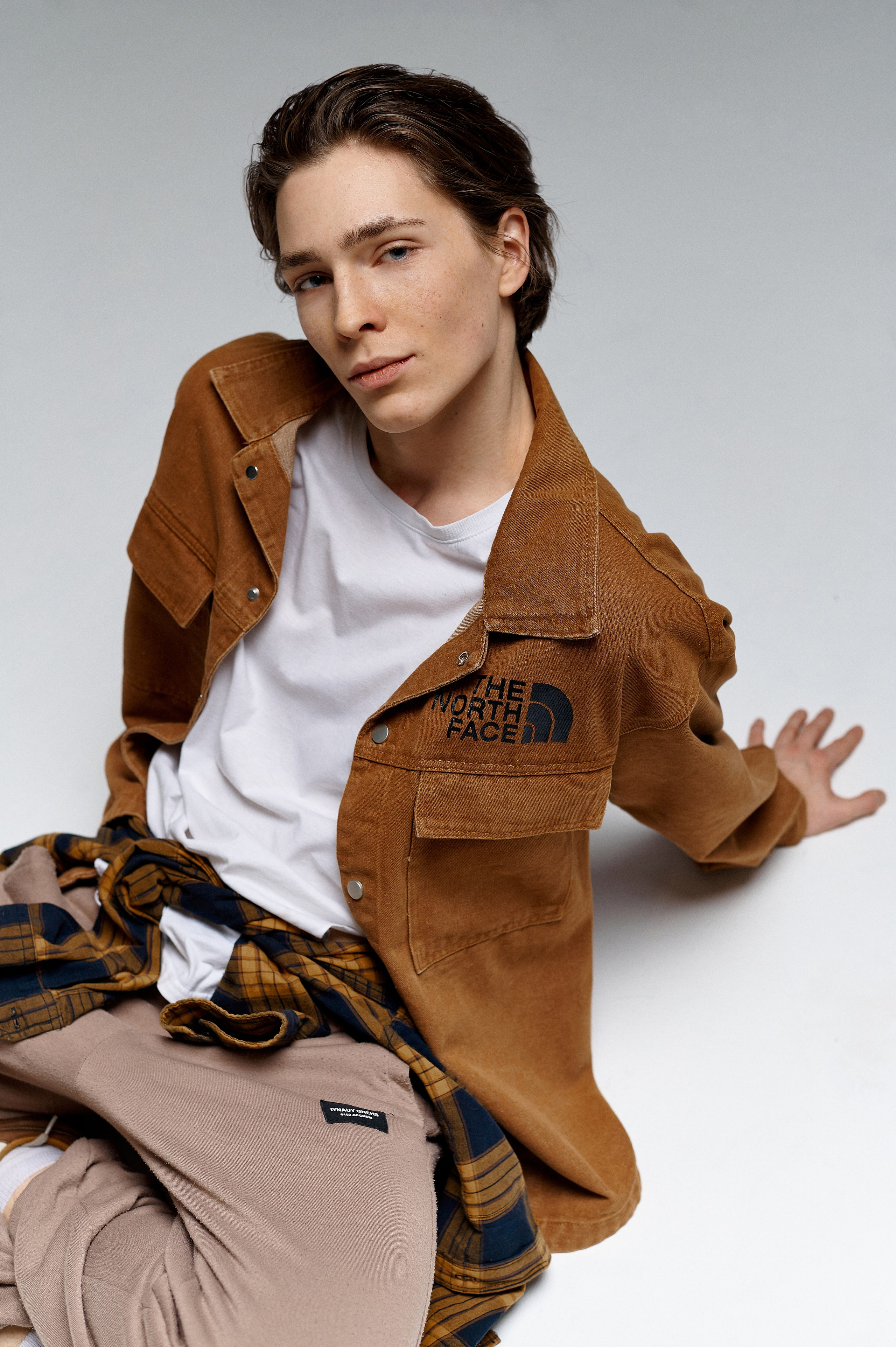 Парень модель в бежевой куртке рубашке и белой футболке лежит на циклораме