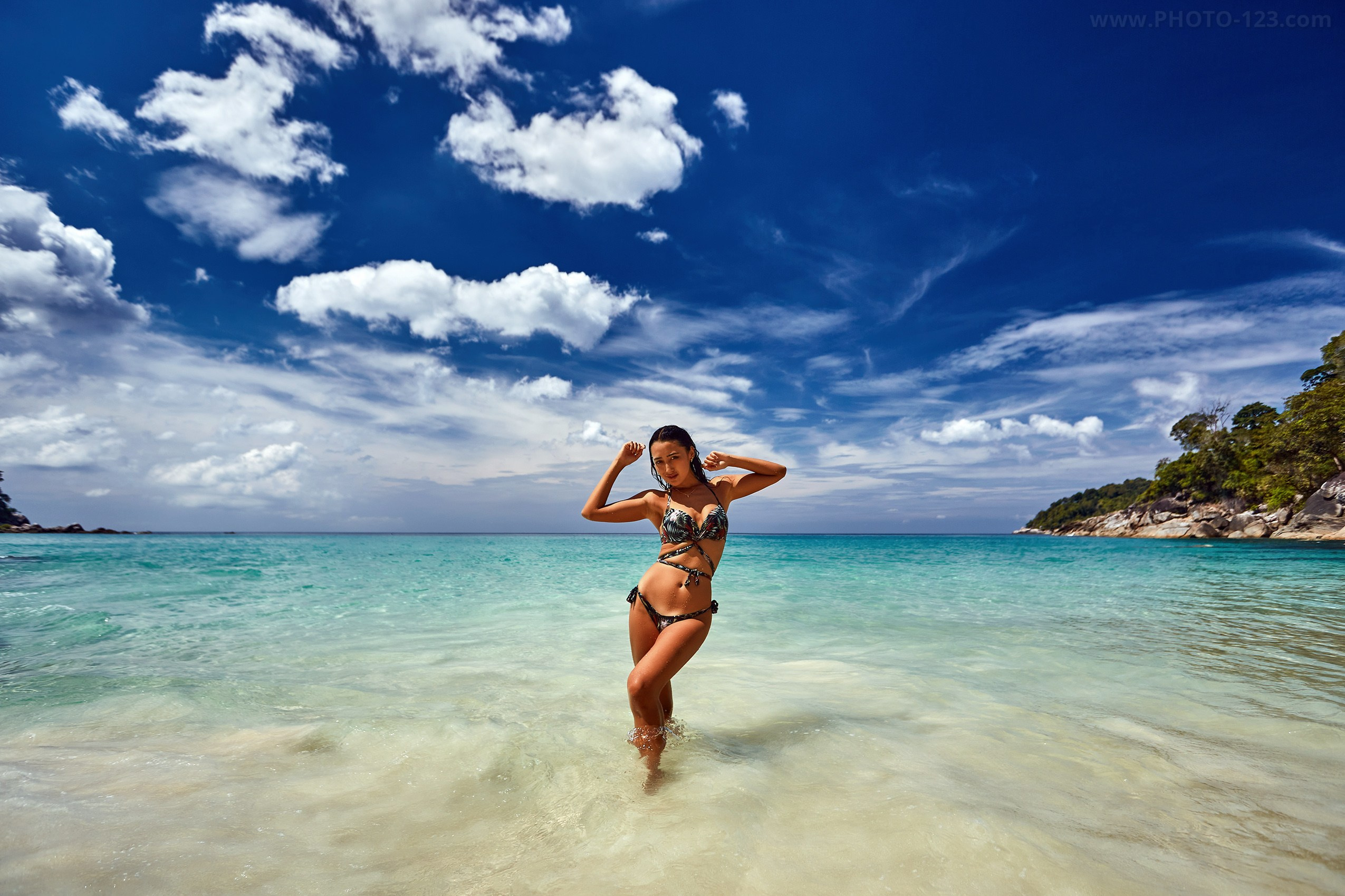 Woman posing in shallow turquoise sea under dramatic blue sky with scattered clouds, tropical beach landscape, bright midday light, photographer in Phu Quoc
