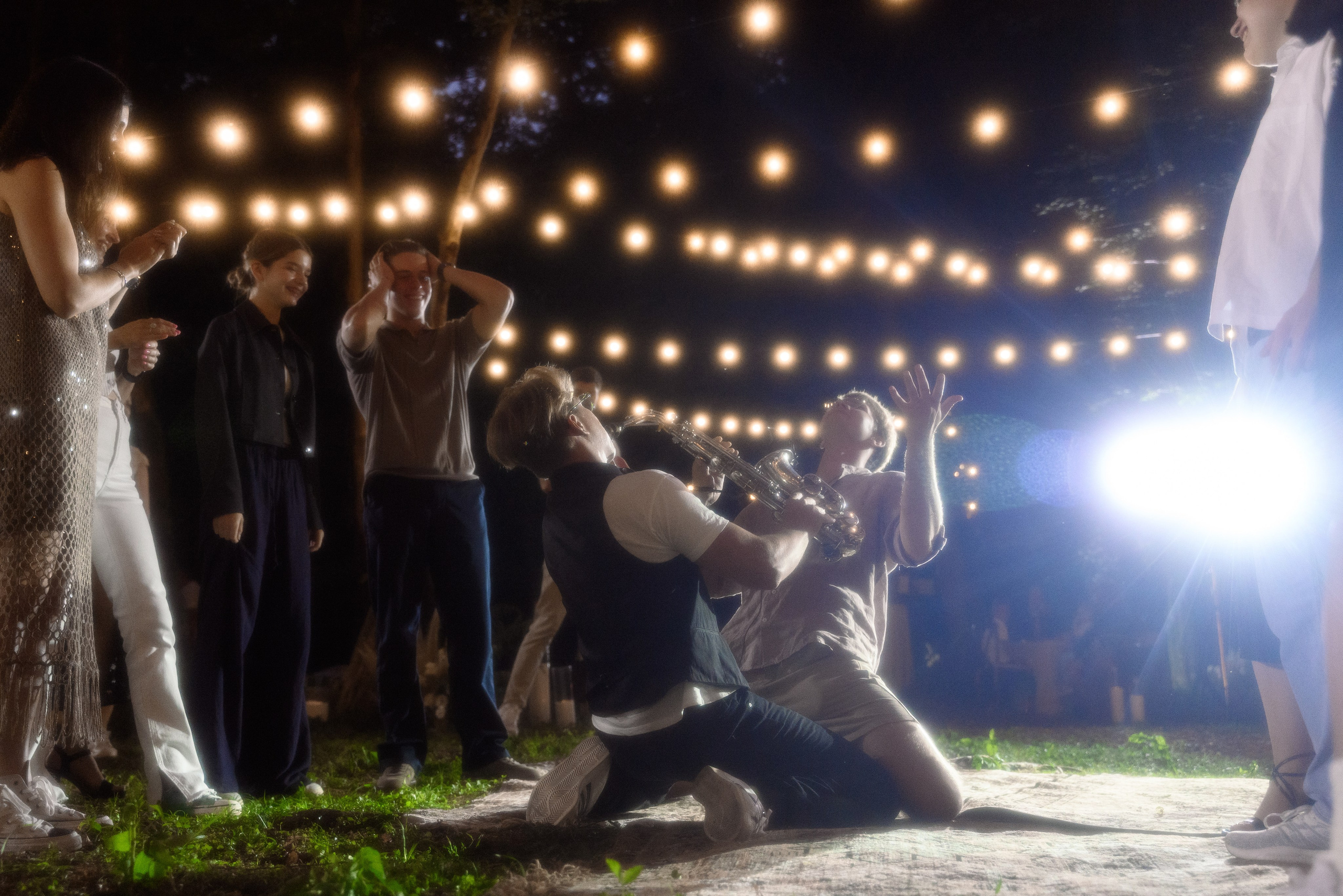 Вечеринка свадебная в лесу люди аплодируют по бокам а в центре кадра саксофонист на коленях играют музыку и рядом с ним также на коленях стоит гость они оба радуются и празднуют