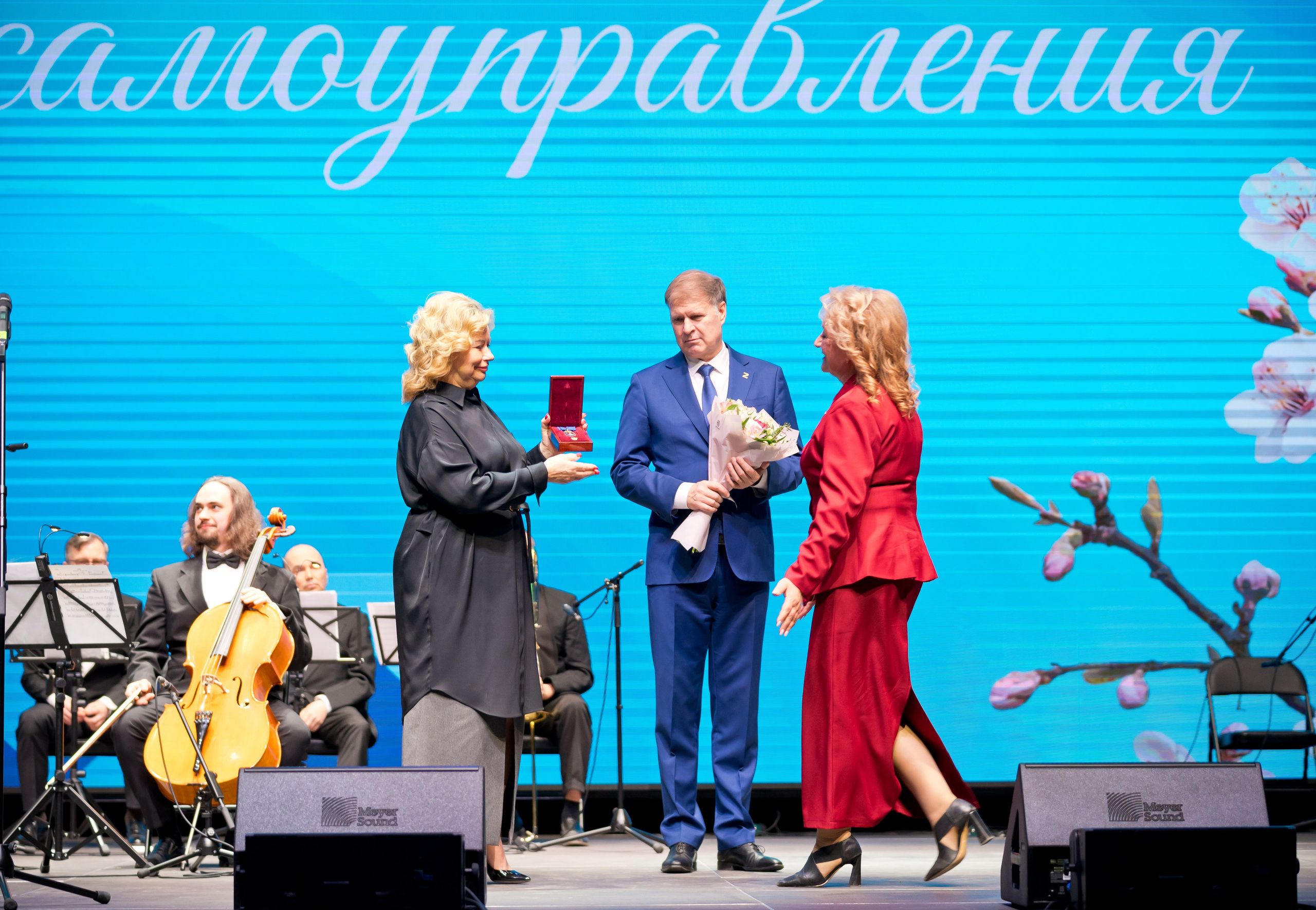 Церемония награждения: женщина в чёрном вручает красную коробочку с наградой, мужчина в синем костюме держит букет. День местного самоуправления. Фото Евгения Мальцев.