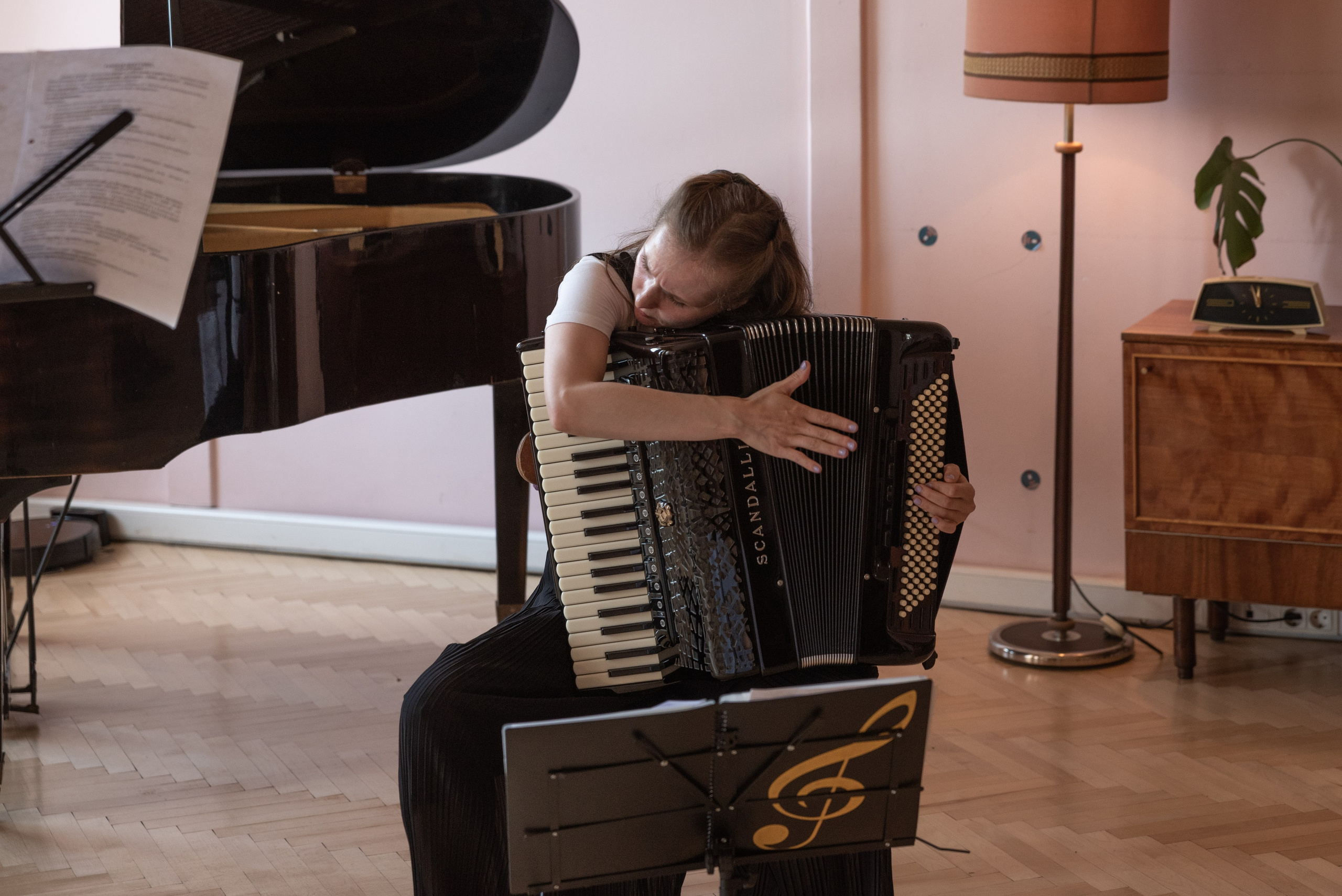 Девушка с аккордеоном сидит на стуле, положила голову на аккордеон. Рядом стойка с нотами, за спиной рояль.