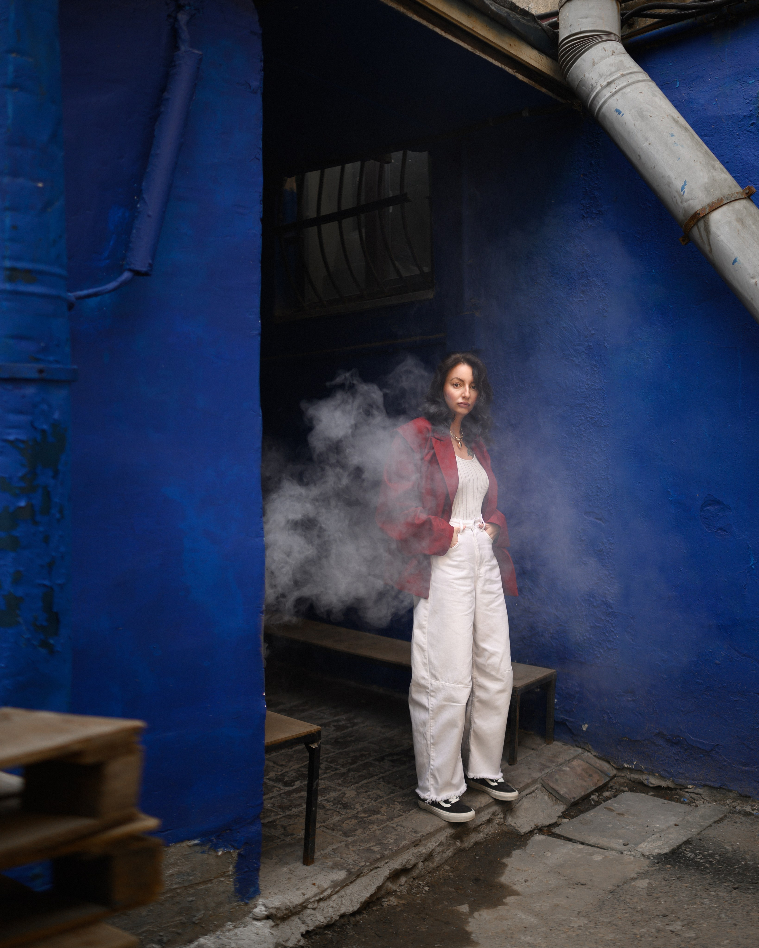 девушка в красном пиджаке позирует на фоне синей стены в дыме и тумане
