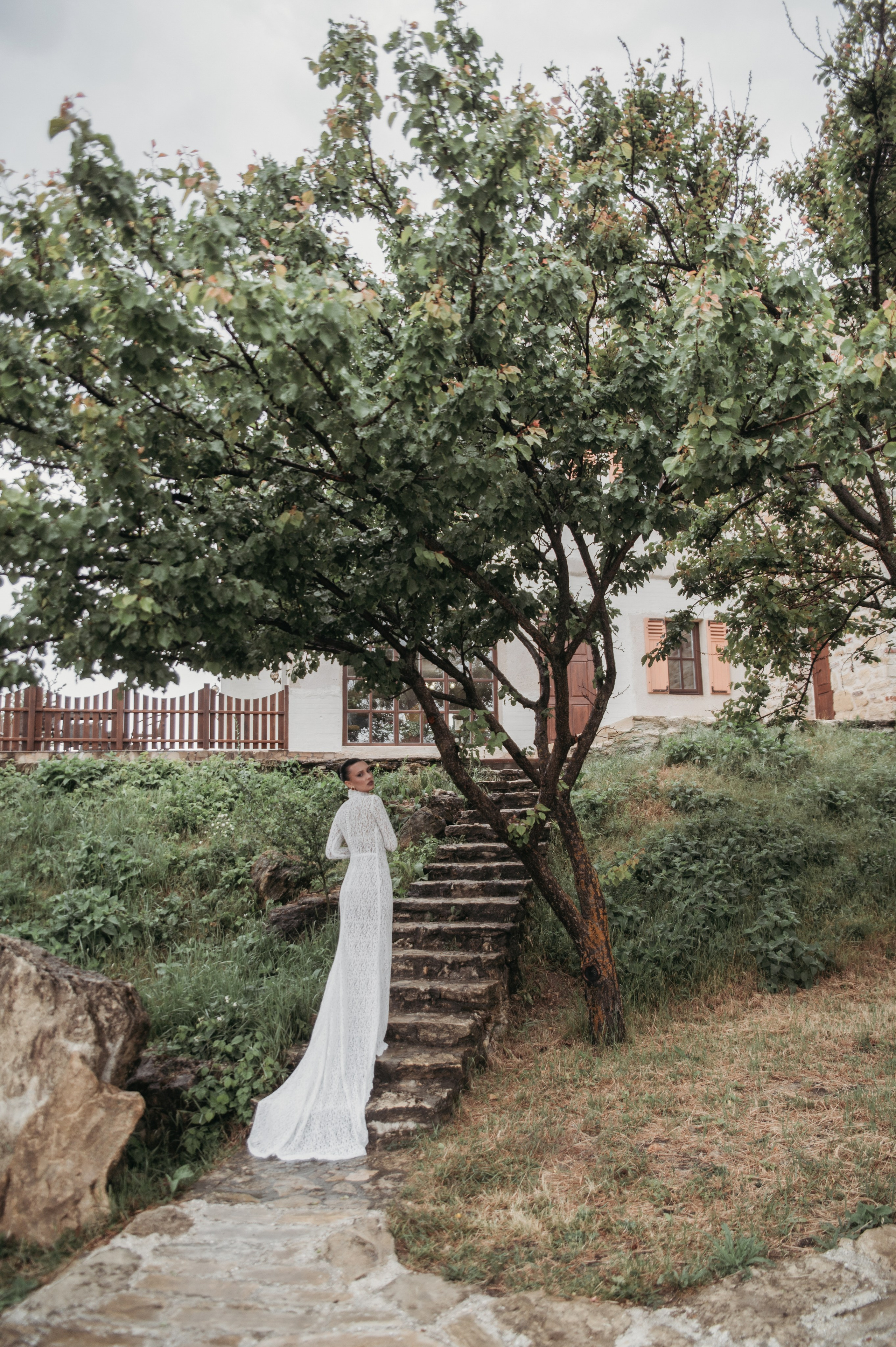Невеста в длинном свадебном платье стоит у ступеней, окруженная зелеными деревьями, создавая атмосферу романтики и естественной красоты