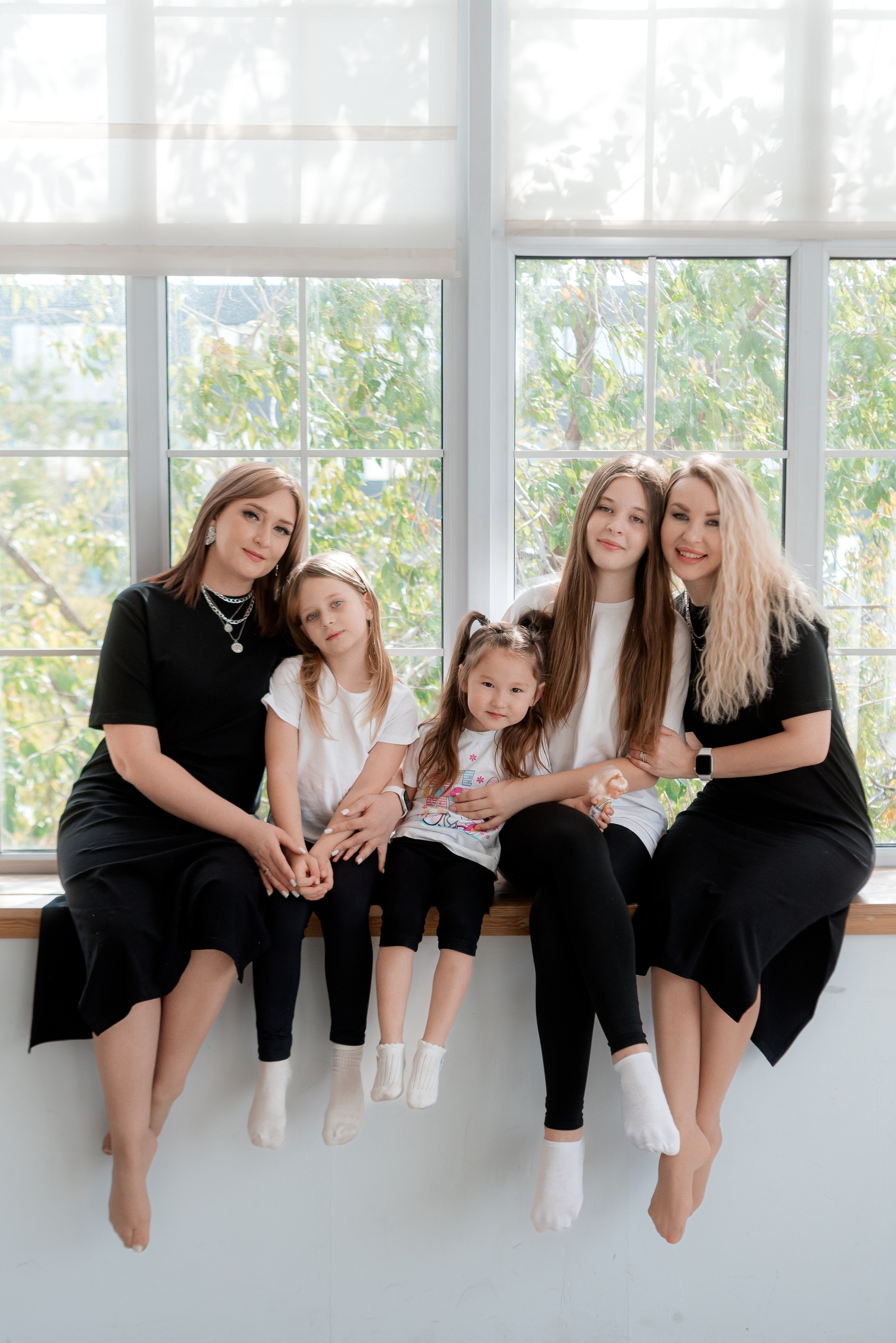 Две подруги со своими дочерьми сидят на подоконнике и все вместе обнимаются