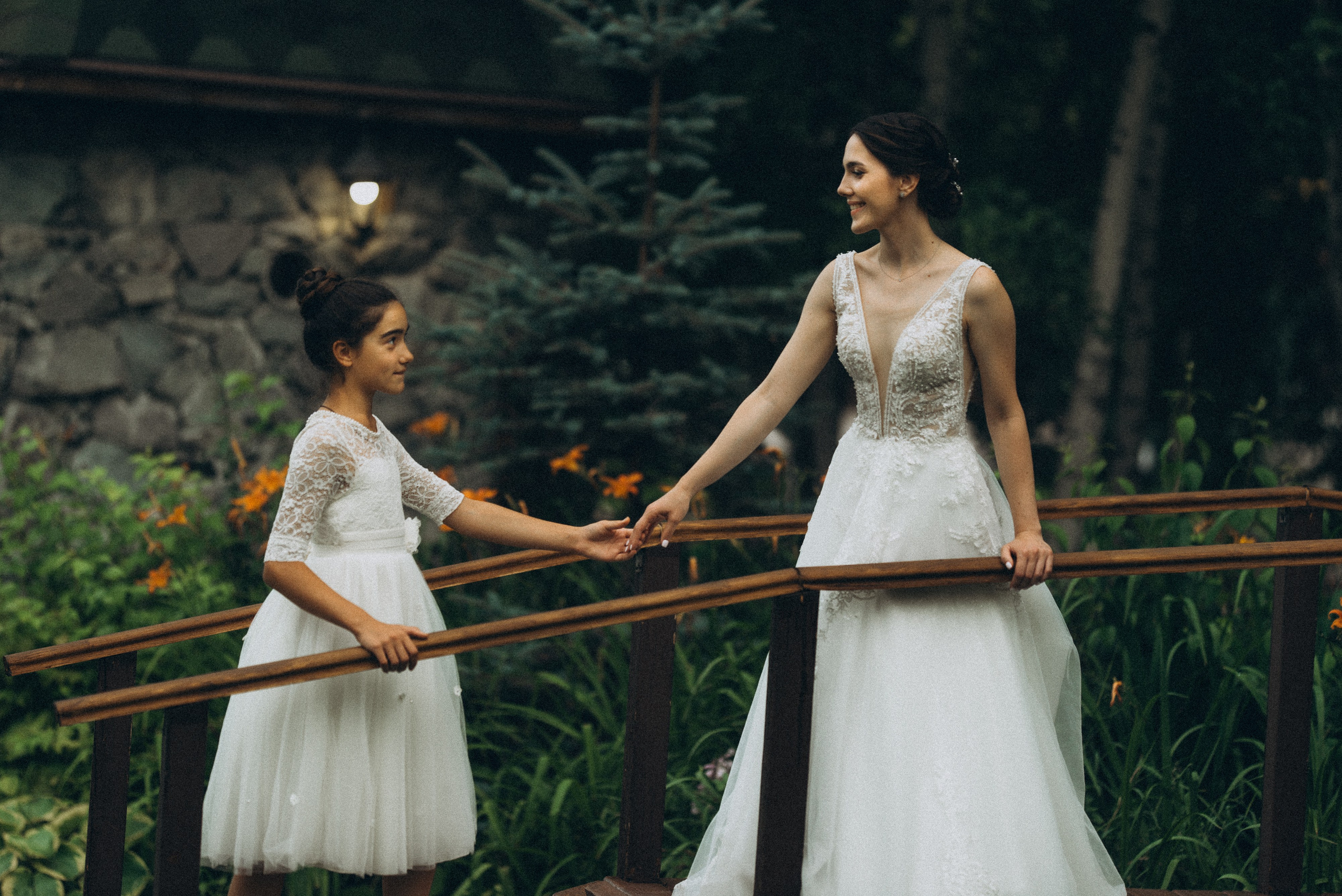 Невеста Линда и маленькая девочка в белом платье держатся за руки на деревянном мостике. Свадебная фотосессия с детьми.