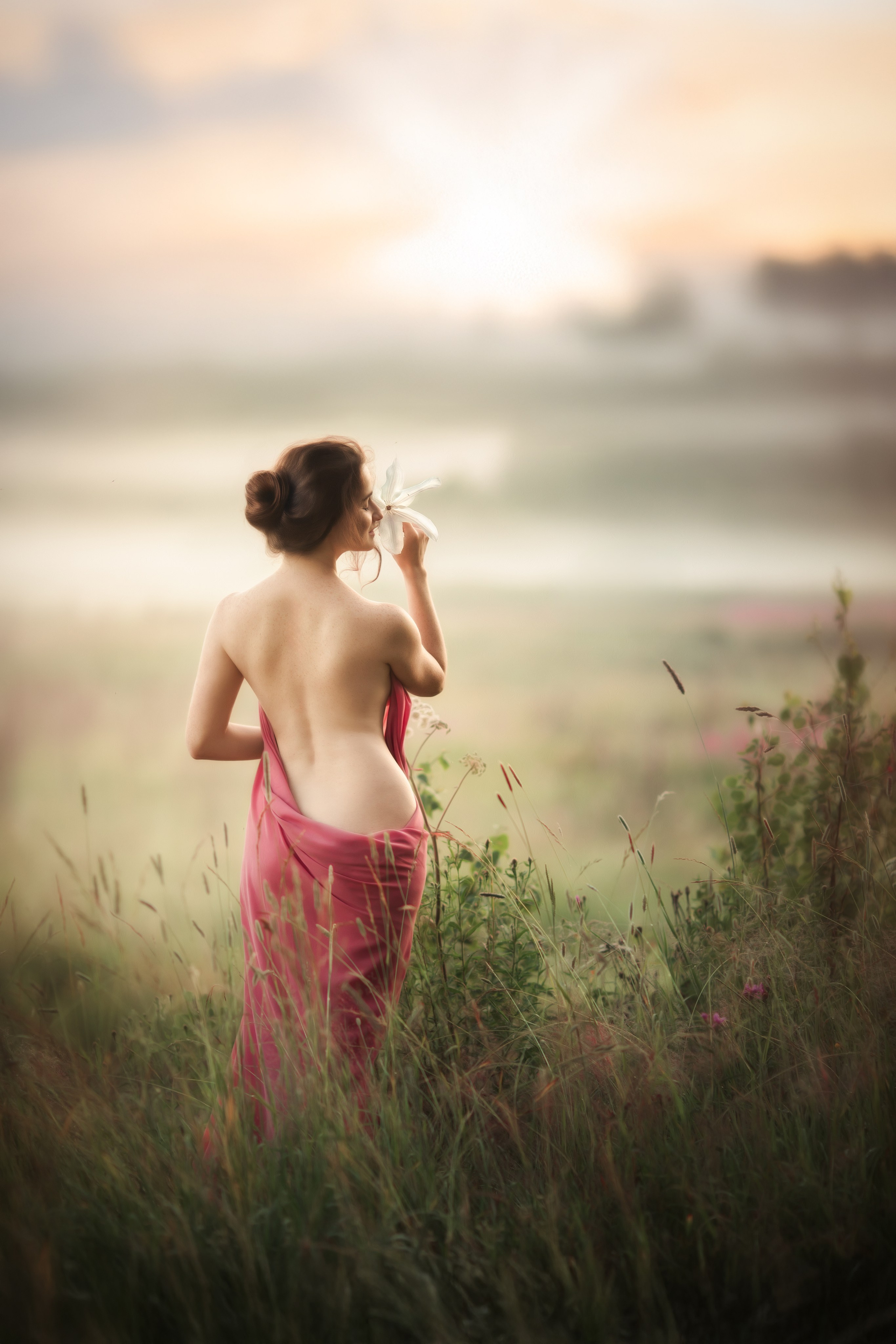 Женщина в розовой ткани стоит спиной в поле на рассвете, держит белый цветок у лица, фон залит мягким светом и туманом.
