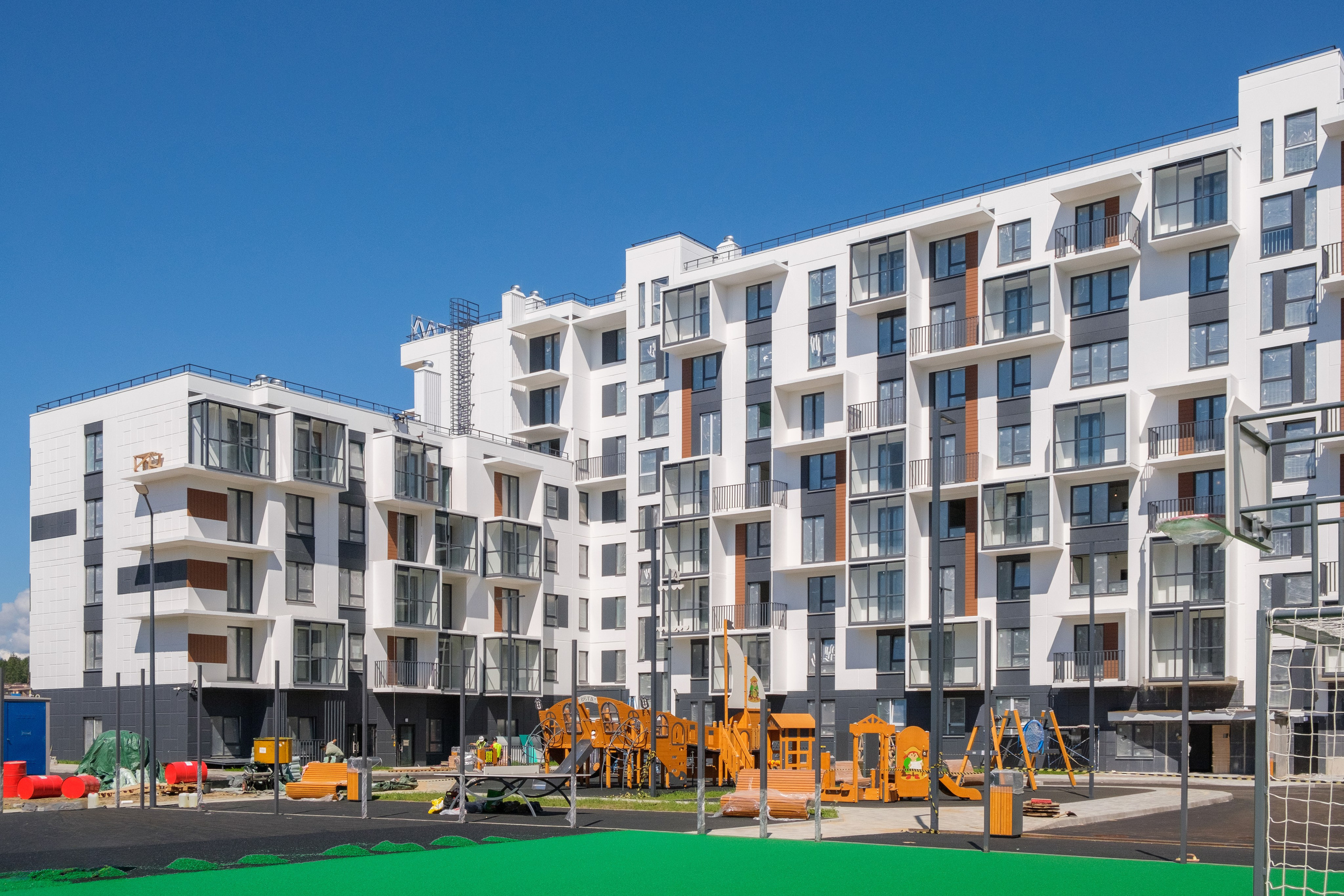 На изображении показаны несколько зданий современной архитектуры, вероятно, это жилой комплекс. Здания имеют белый и серый цвета, окна расположены таким образом, чтобы обеспечить максимальное естественное освещение внутри помещений. Вокруг зданий видны строительные материалы и оборудование, что указывает на то, что строительство ещё продолжается.