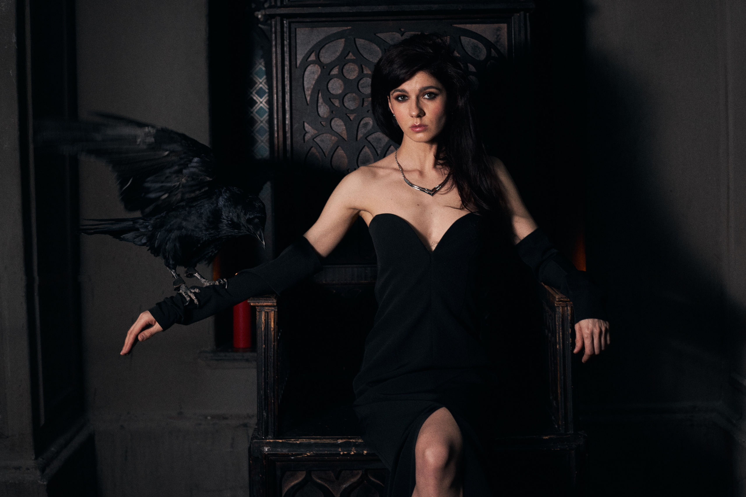 Красавица-брюнетка с длинными волосами в открытом черном платье сидит на троне и держит на руке ворона
