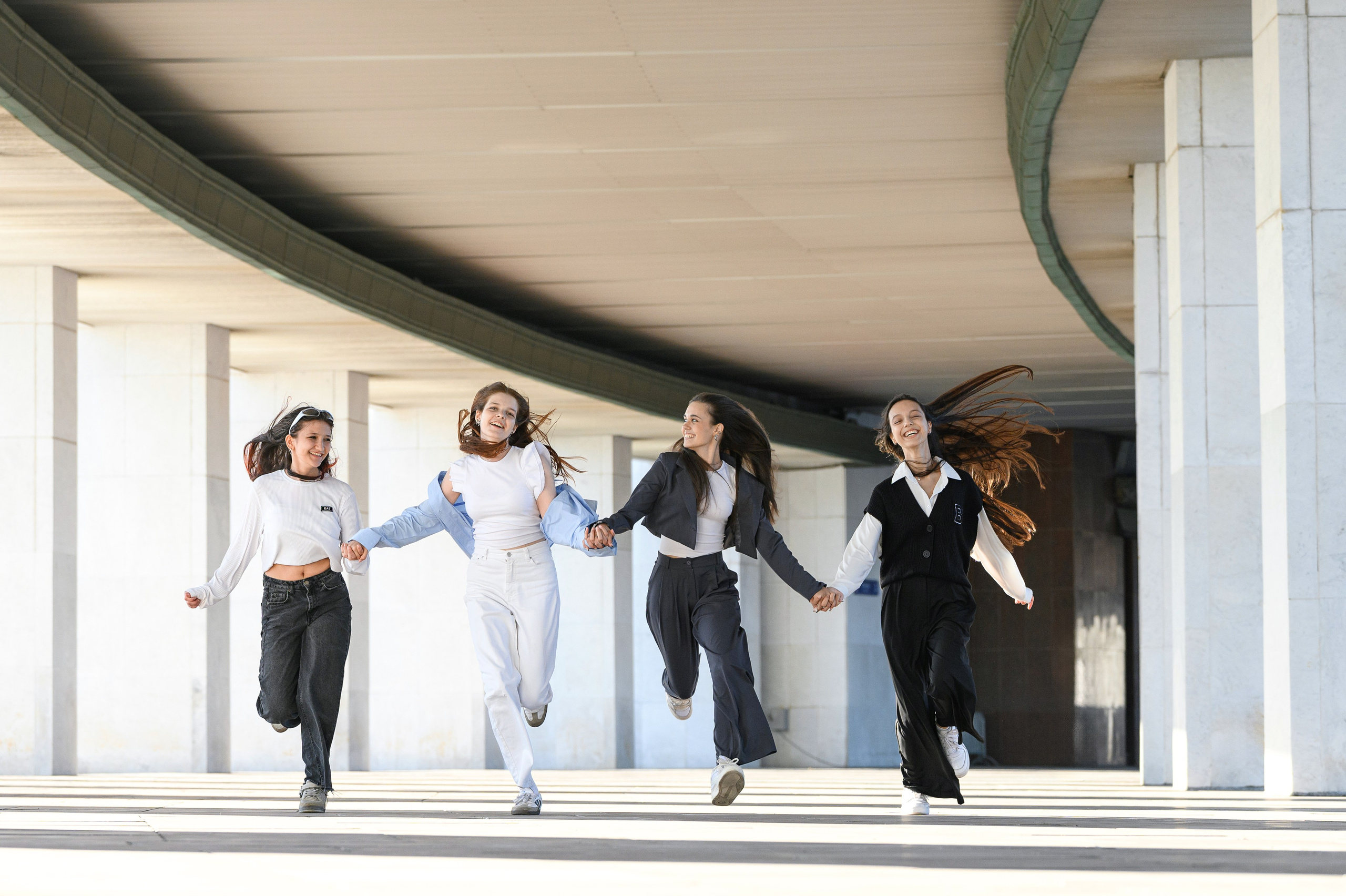 Четверо старшеклассниц бегут по галерее с колоннами в динамичном кадре выпускной фотосессии.