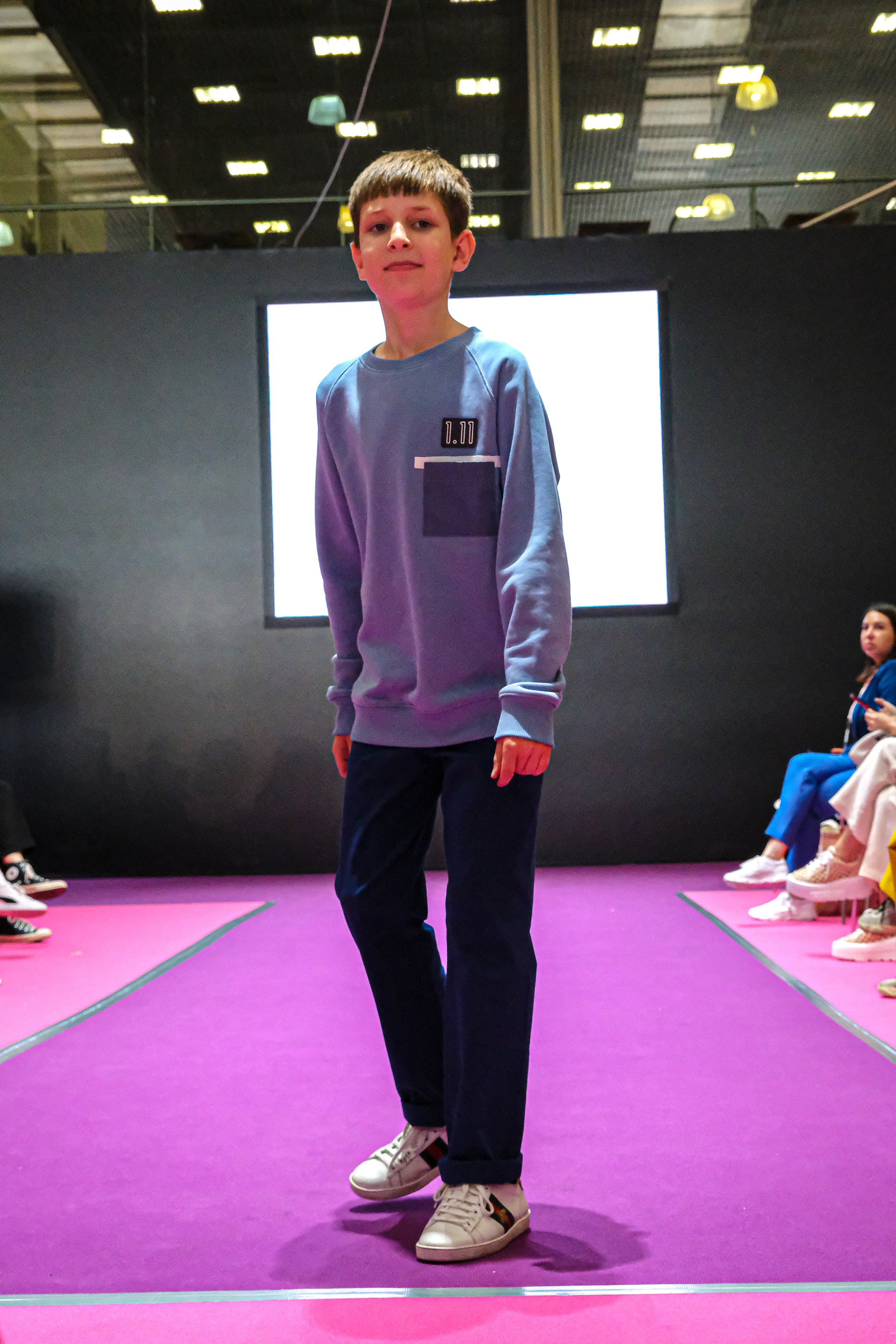  "Модели в ярких нарядах проходят по подиуму во время показа мод, освещенные яркими софитами и окруженные восторженными зрителями."