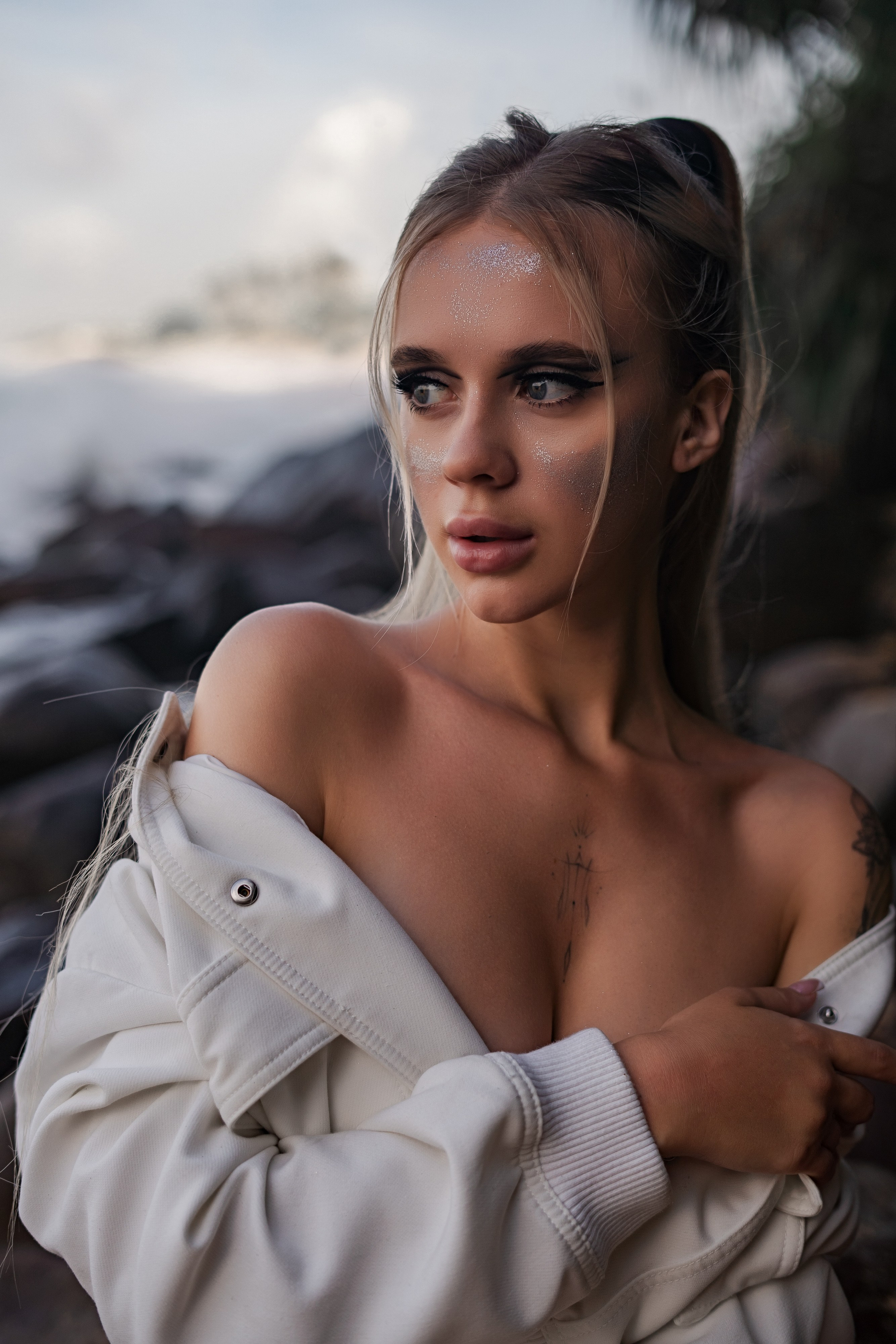 Very beautiful blonde with makeup on the rocks by the ocean in a white jacket
