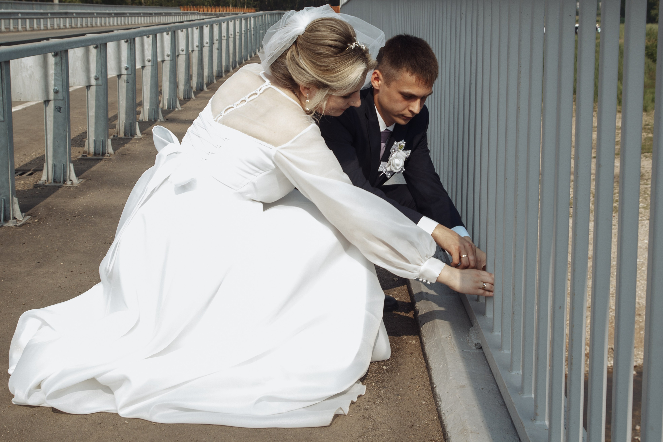 Невеста помогает жениху с обувью, сидя с ним на перилах моста — трогательный момент заботы.