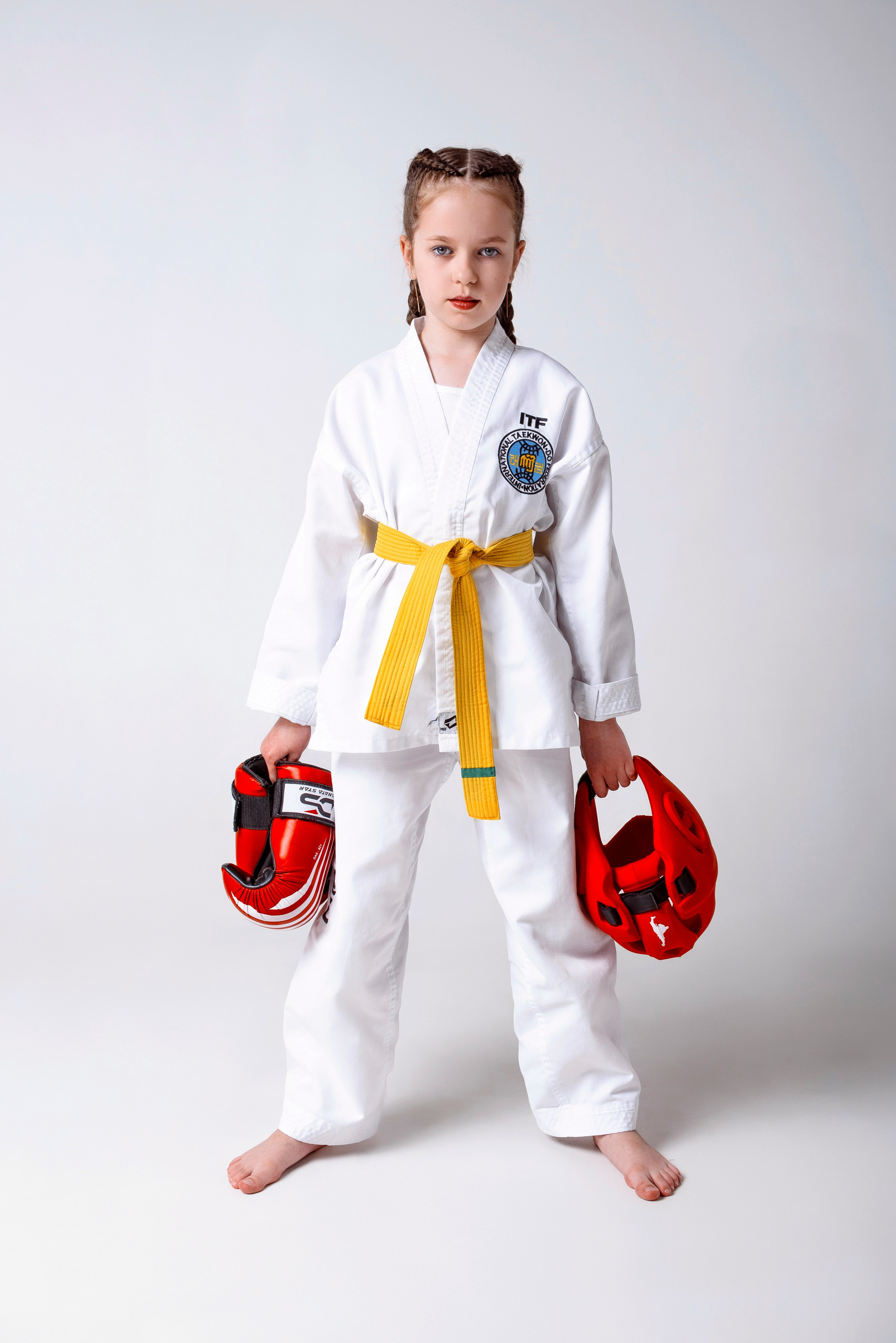 На изображении изображено лицо, одетое в белую форму Тхэквондо (добок) с желтым поясом. Человек носит красные боксерские перчатки и, вероятно, готовится к тренировке боевых искусств. На форме есть нашивки, одна из которых содержит буквы "ITF", что, вероятно, указывает на Международную федерацию Тхэквондо. Фон светлый и однородный, что обеспечивает четкий обзор объекта.