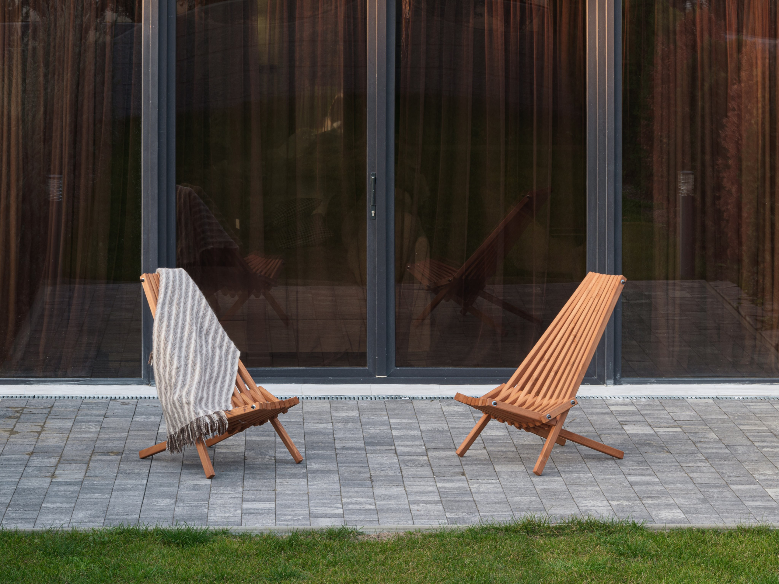 Две деревянные шезлонг-кресла на террасе частного дома, одно из которых украшено мягким вязаным пледом в бежевых тонах, на фоне панорамных стеклянных дверей. Атмосфера уюта и минимализма подчеркнута дизайном Алины Дёровой, стилист Наталья Якушева, фото Сергей Денисенко.