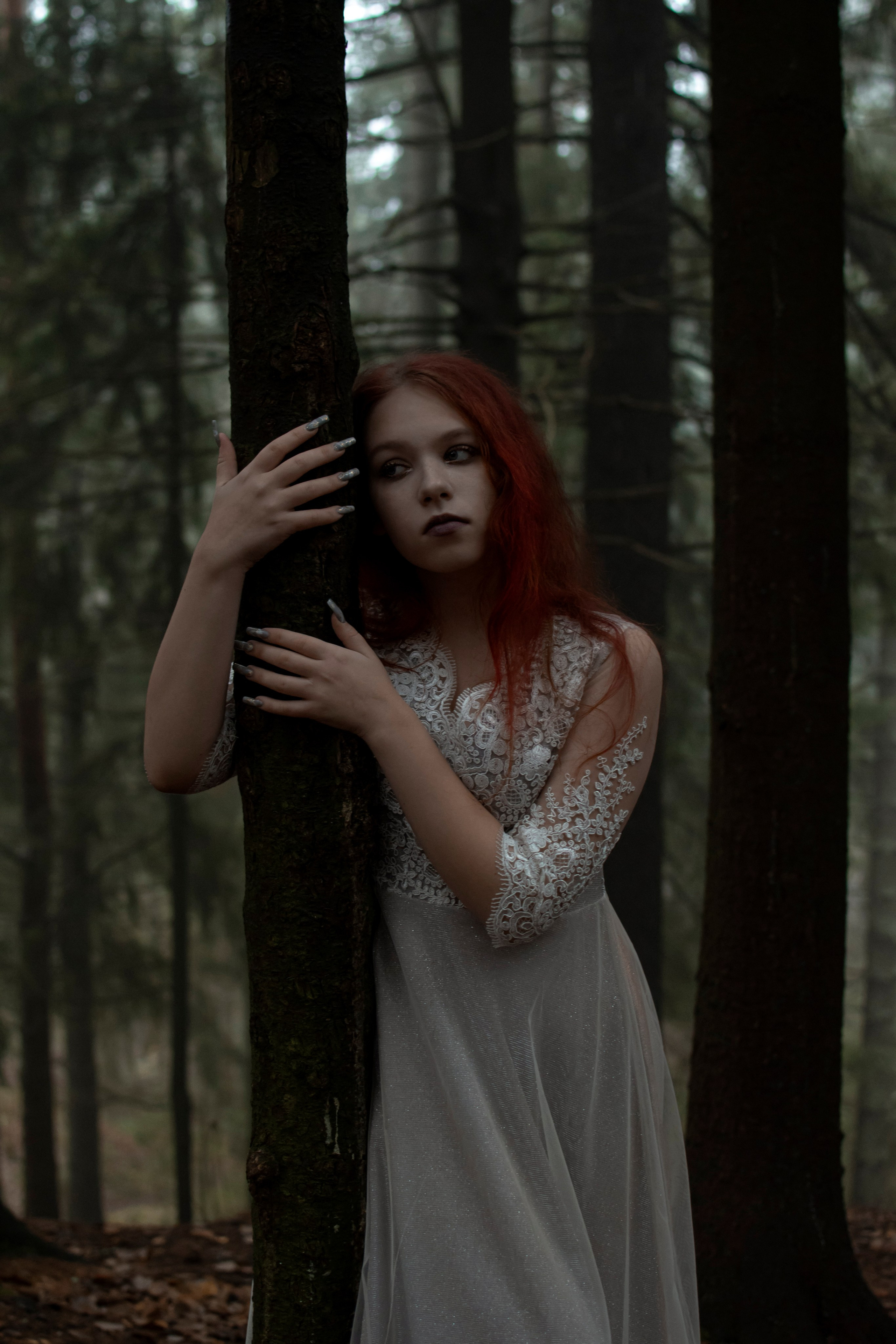 Красноволосая девушка в белом платье обнимает сосну в лесу