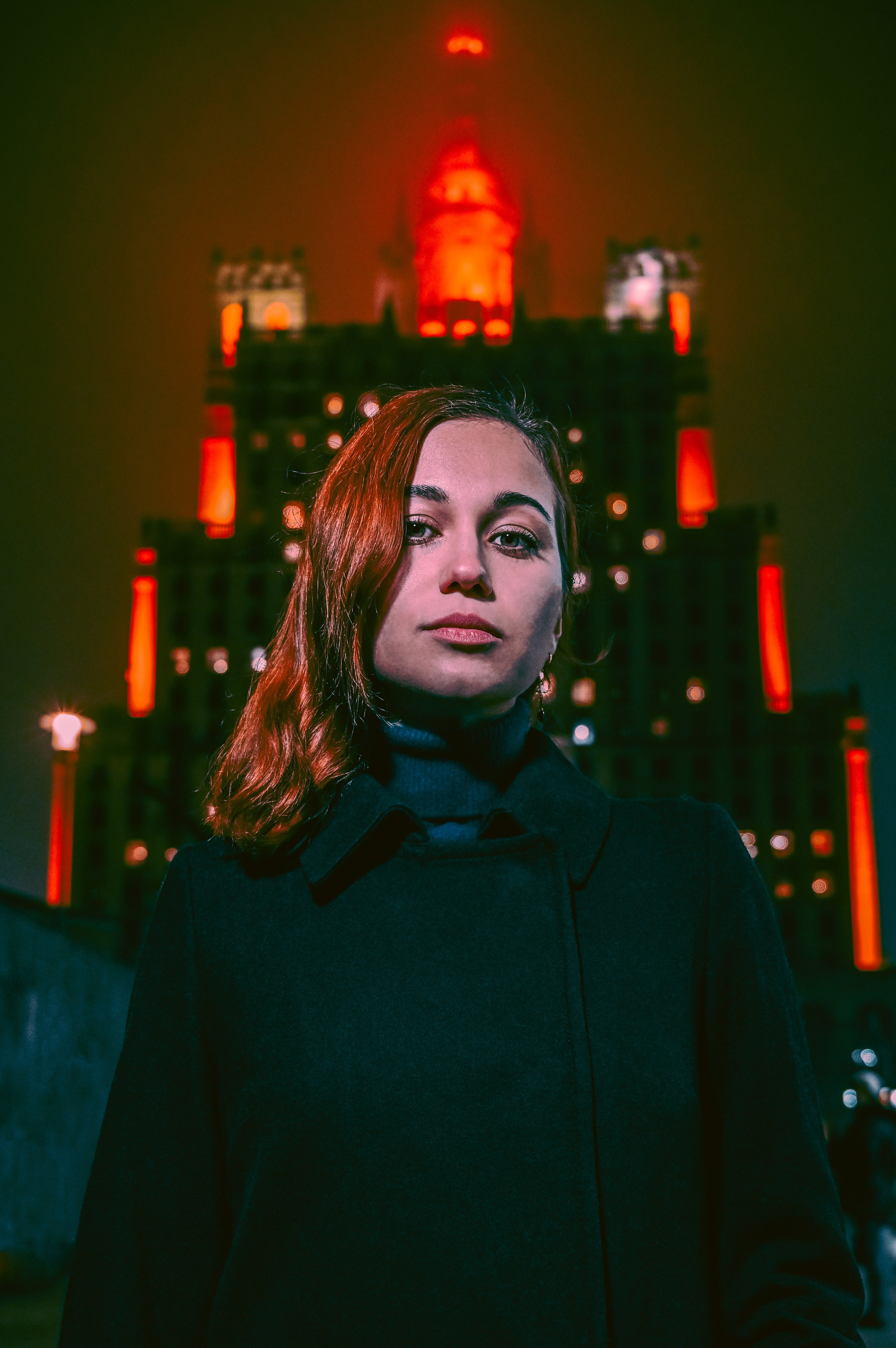 Ночной портрет женщины с волнистыми рыжими волосами, снятый в Москве перед архитектурным зданием с красной подсветкой.