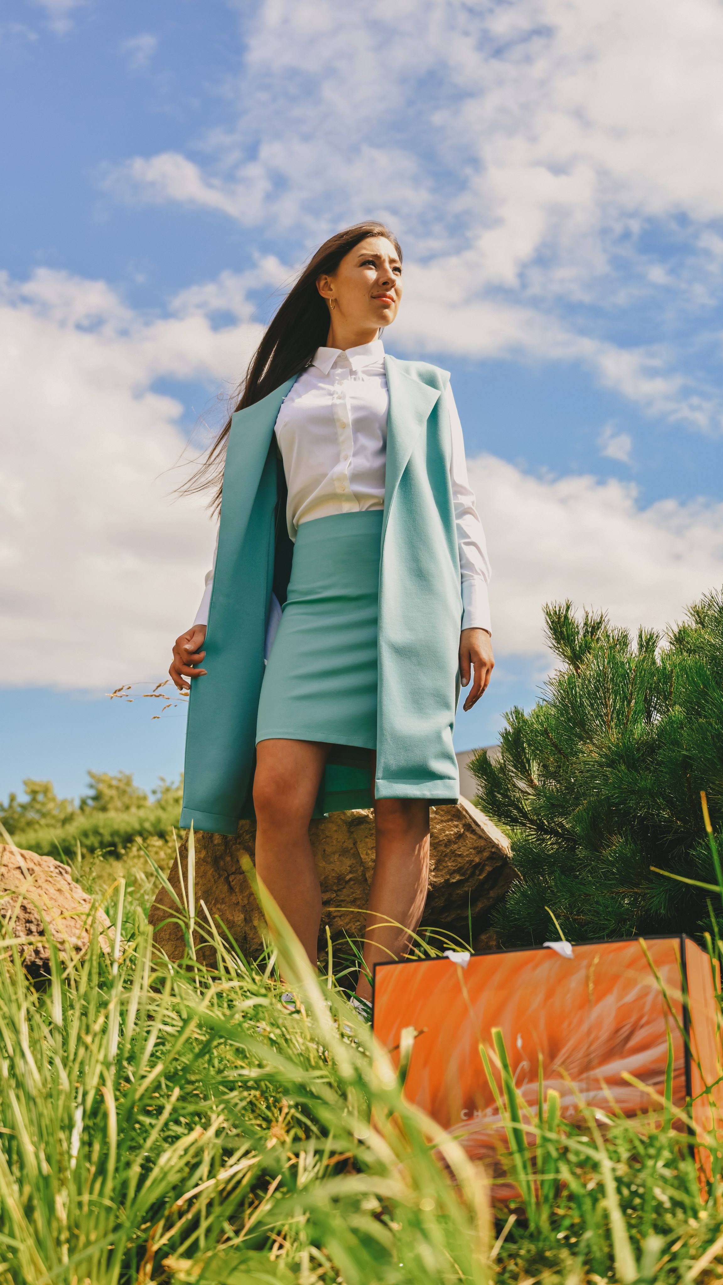 Молодая женщина в элегантном образе с белой блузкой, светло-зеленой юбкой и жакетом стоит на фоне живописного летнего пейзажа. Она одета в стильный комплект, который идеально сочетается с природной обстановкой. Волосы аккуратно собраны назад, а ее взгляд направлен в сторону, создавая ощущение задумчивости и спокойствия. На переднем плане видны высокие травы и камни, что добавляет фотографии естественную текстуру и глубину. Светлый фон неба с легкими облаками подчеркивает свежесть и чистоту момента. Этот снимок демонстрирует профессиональное владение техникой освещения и композиции, делая акцент на игре света и тени, а также на гармоничном сочетании человека и природы. Perfect for fashion and outdoor photography enthusiasts!