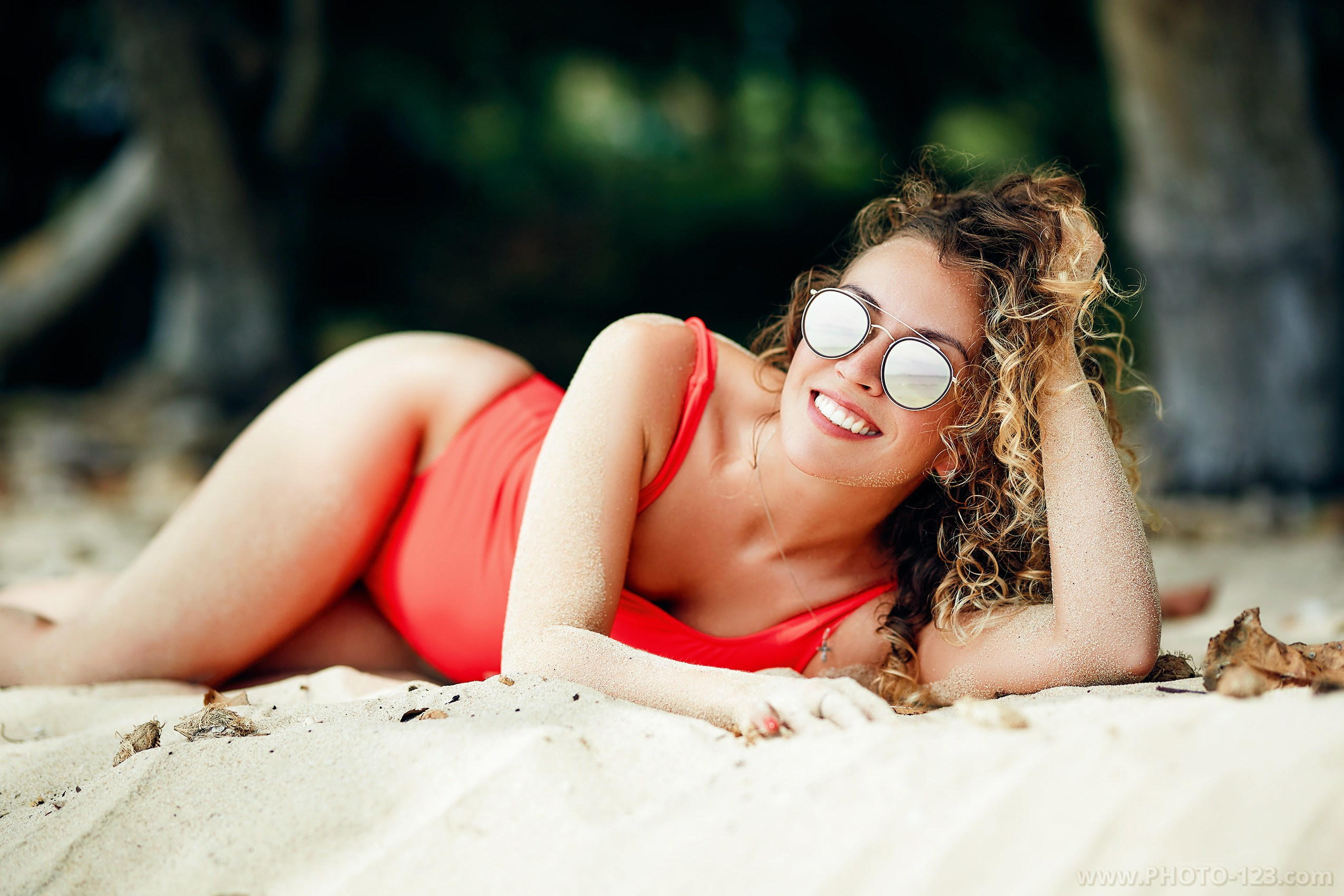 Lifestyle portrait of a woman in red swimsuit lying on sandy beach with curly hair and reflective sunglasses; smiling with hand in hair, surrounded by tropical greenery and soft natural light; editorial-style image captured in Phu Quoc