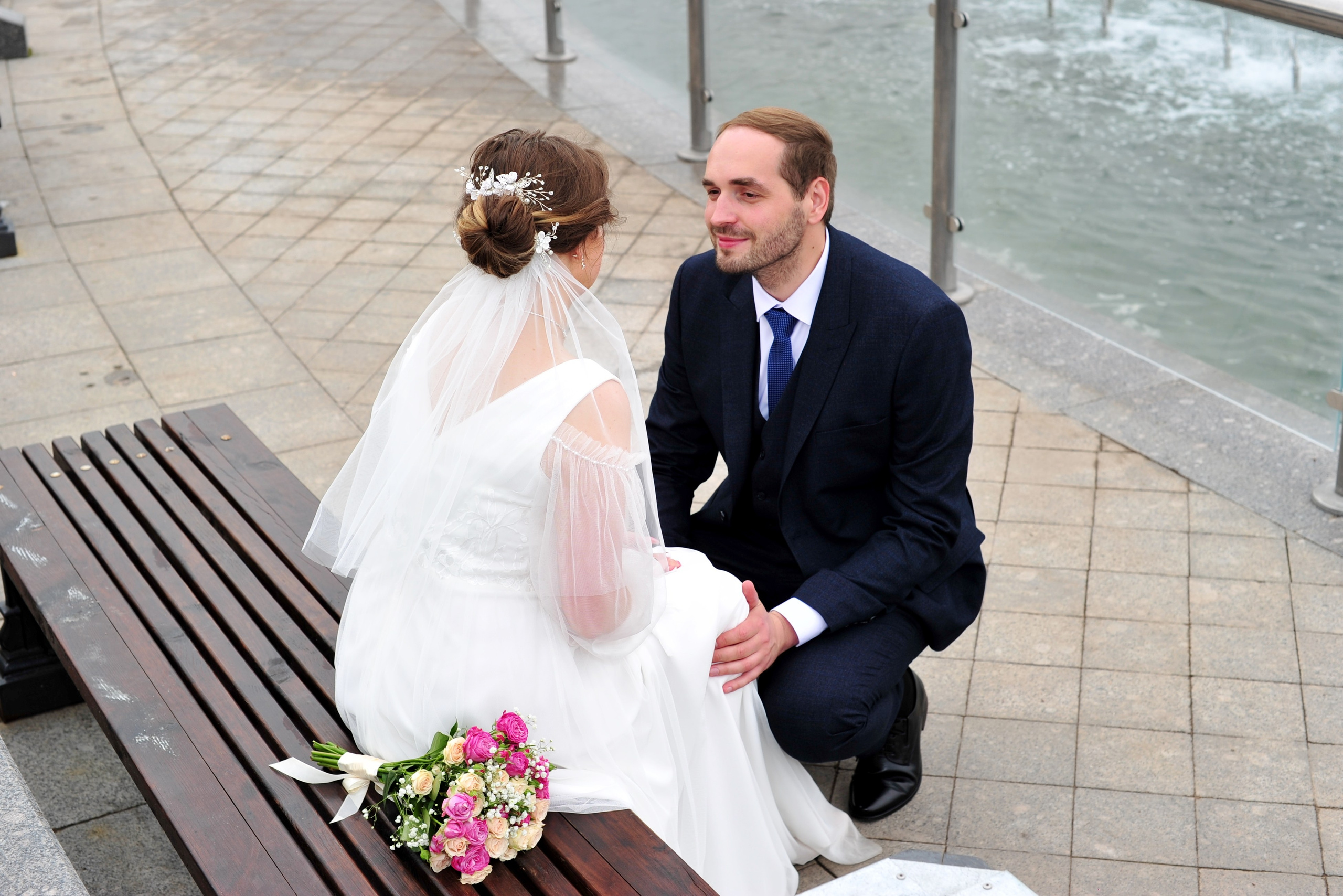 Свадьба в Царицыно пасмурным летним днем, невеста сидит на лавочке у фонтана, жених присел на корточки у ног невесты, смотрит на невесту и  держит ее за колени 