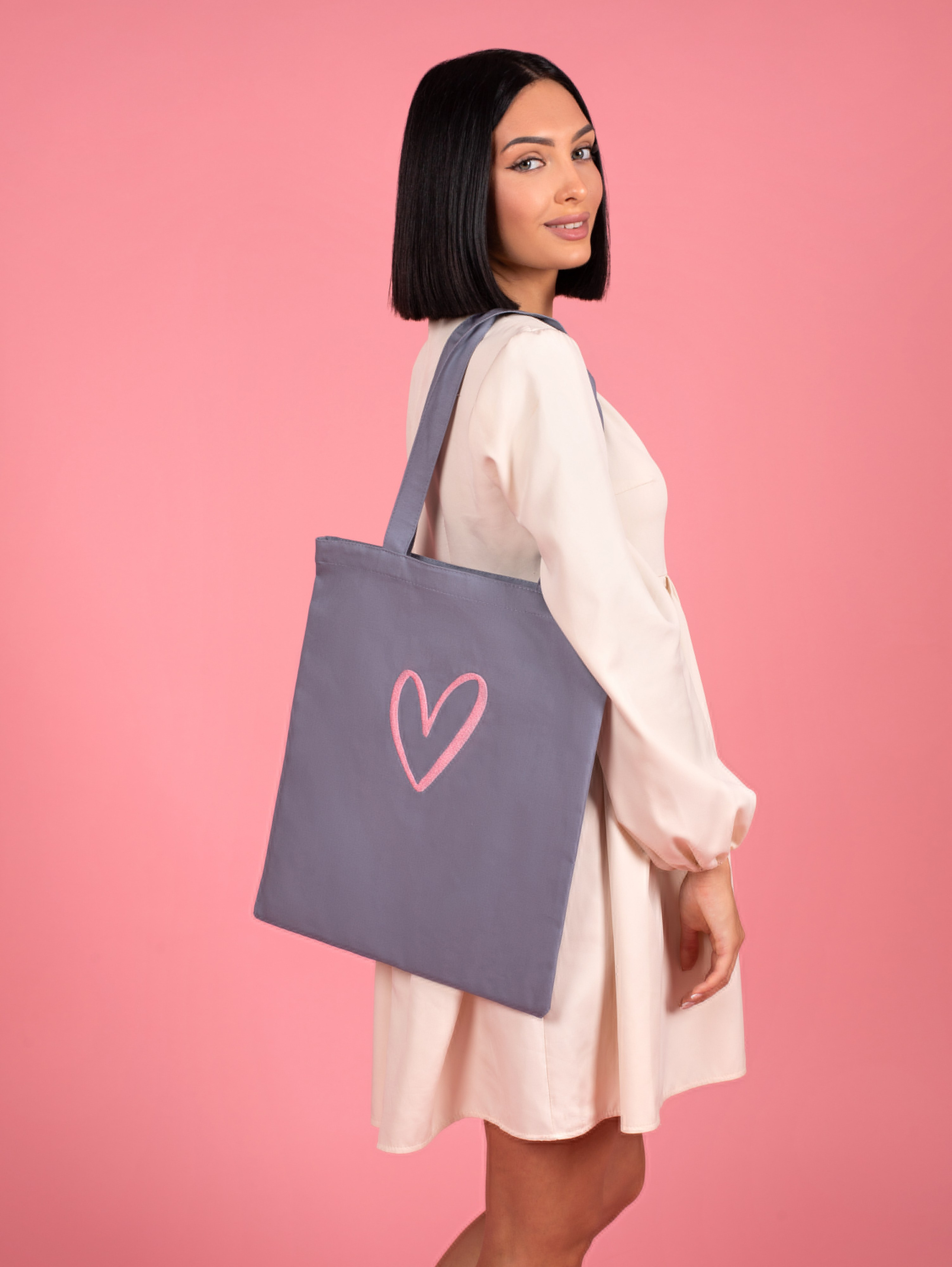 Модель в белом платье и серым шоппером стоит на розовом фоне