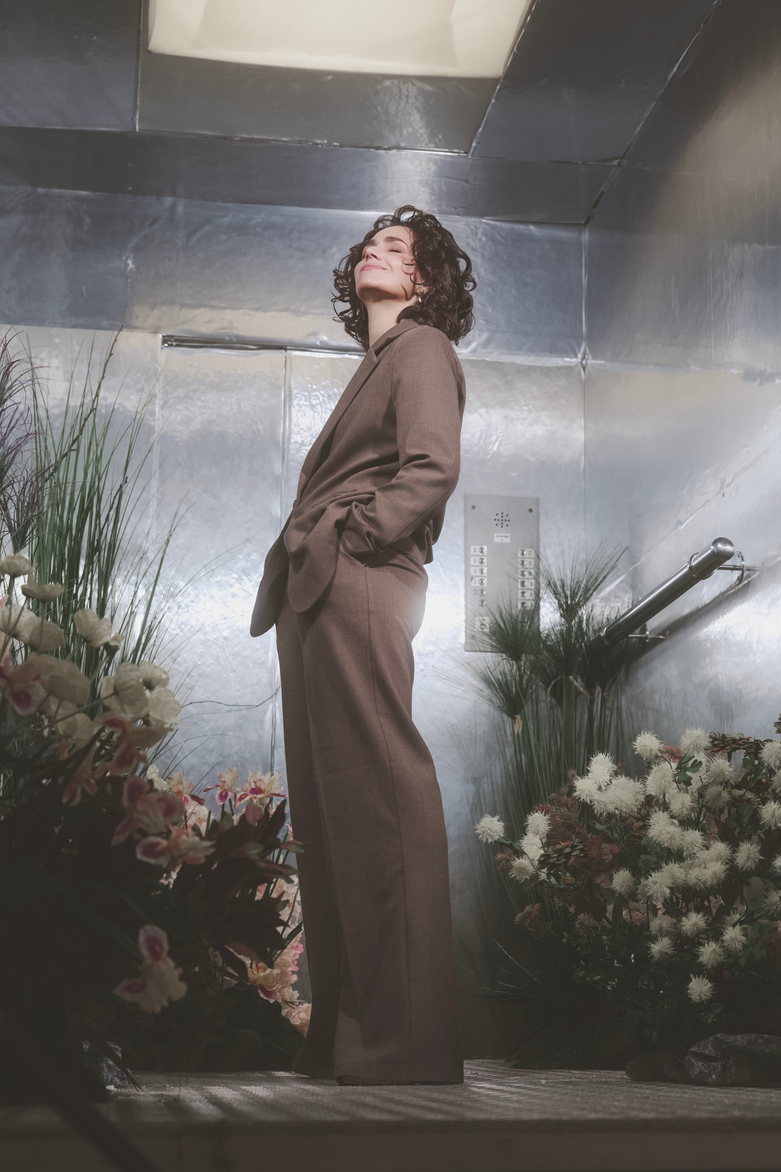 Портретное фото молодой девушки в студии. Девушка сидит на фоне нейтрального цвета, ее лицо освещено мягким светом. Она смотрит прямо в камеру с легкой улыбкой. На девушке стильная одежда, подчеркивающая современный модный стиль. Фотография выполнена в высоком разрешении, с акцентом на деталях