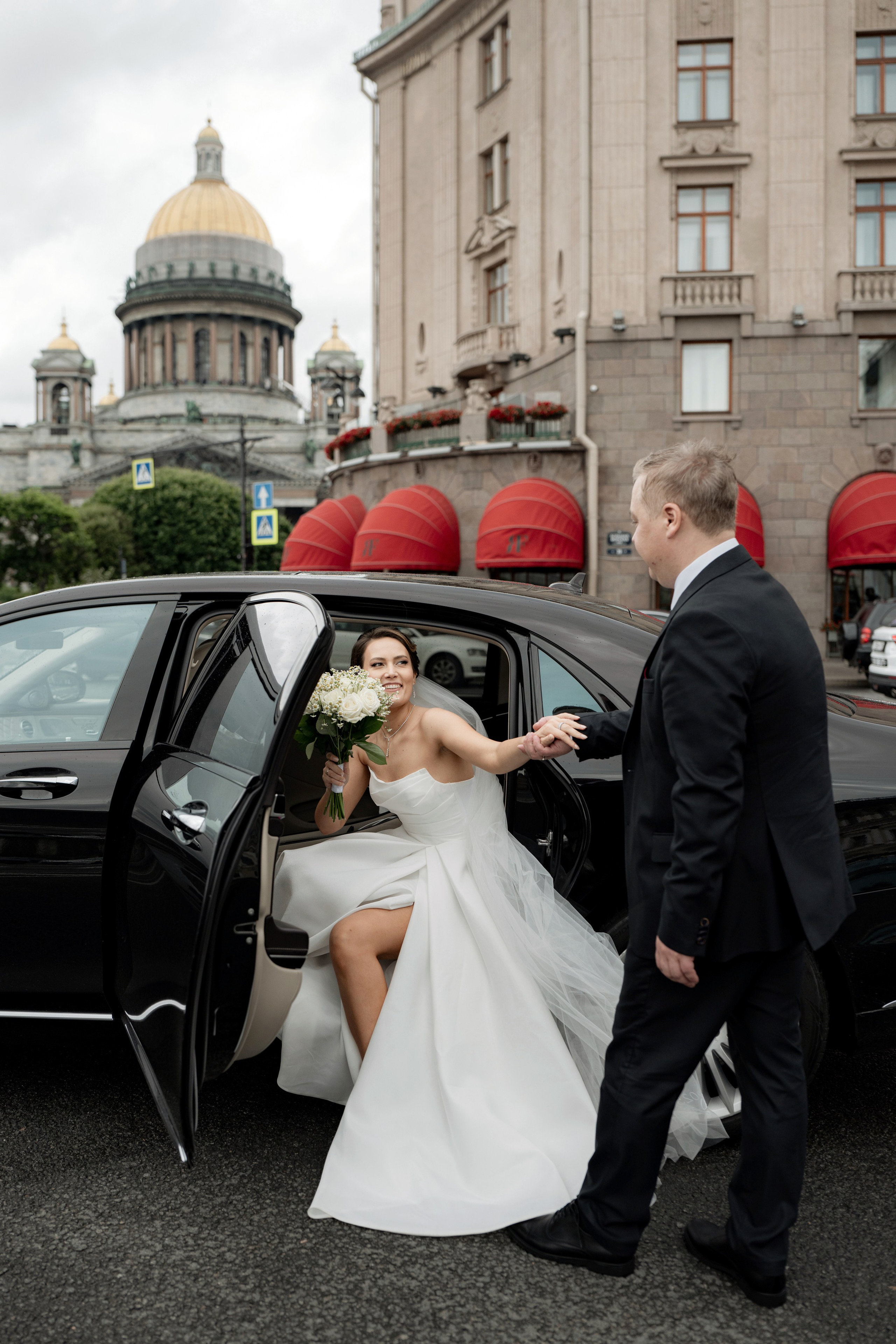 Свадебная фотосессия рядом с Исаакиевским собором. Невеста выходит из черного свадебного автомобиля, жених подает руку на фоне собора и гостиницы Астория. Самые популярные места для свадебной фотосессии в Санкт-Петербурге
