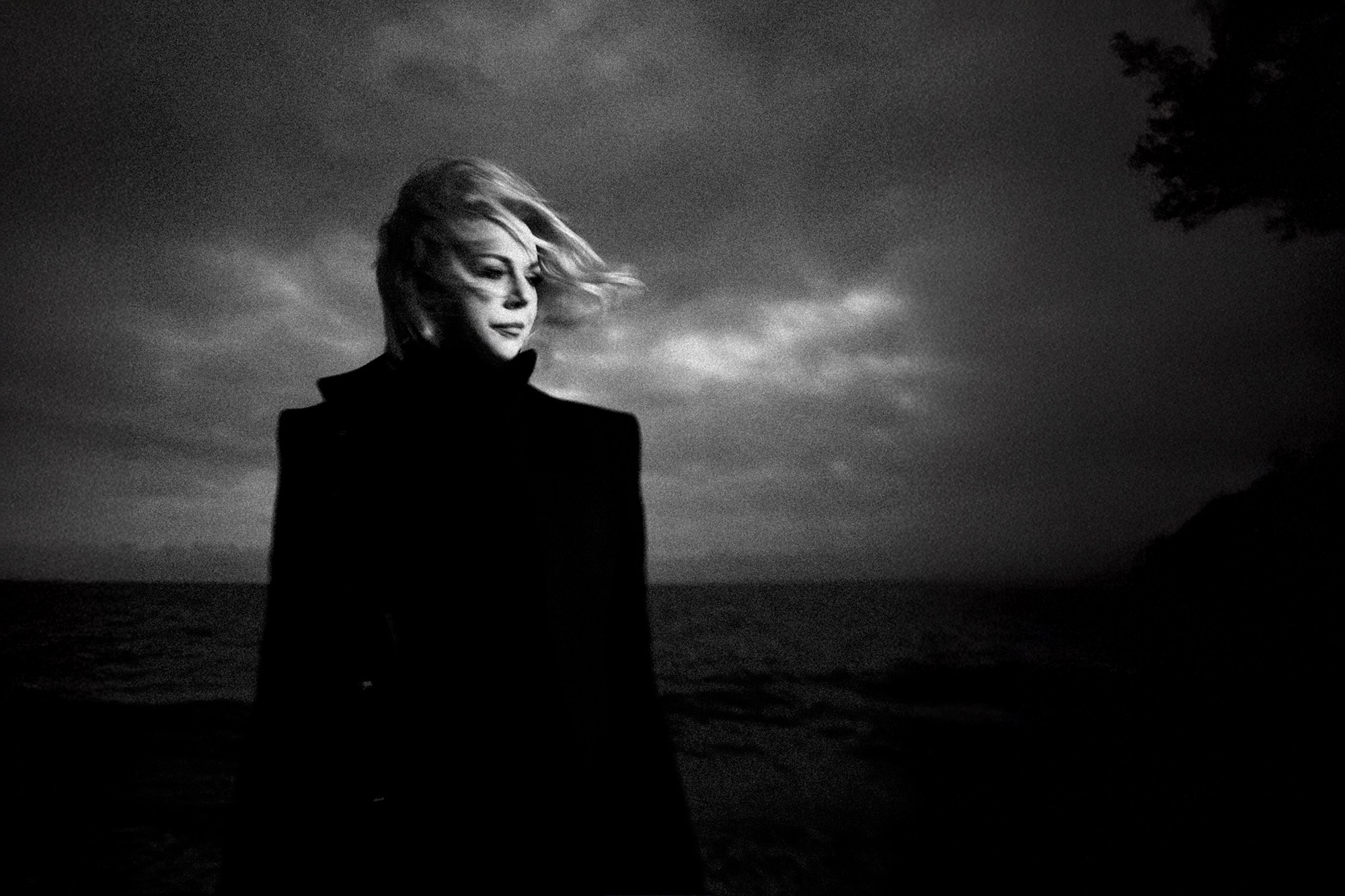 Драматичный черно-белый кадр женщины в темном пальто на фоне штормового моря и пасмурного неба. Ветер развевает волосы, создавая меланхоличное и сильное настроение. Портретный фотограф Галина Скорик.