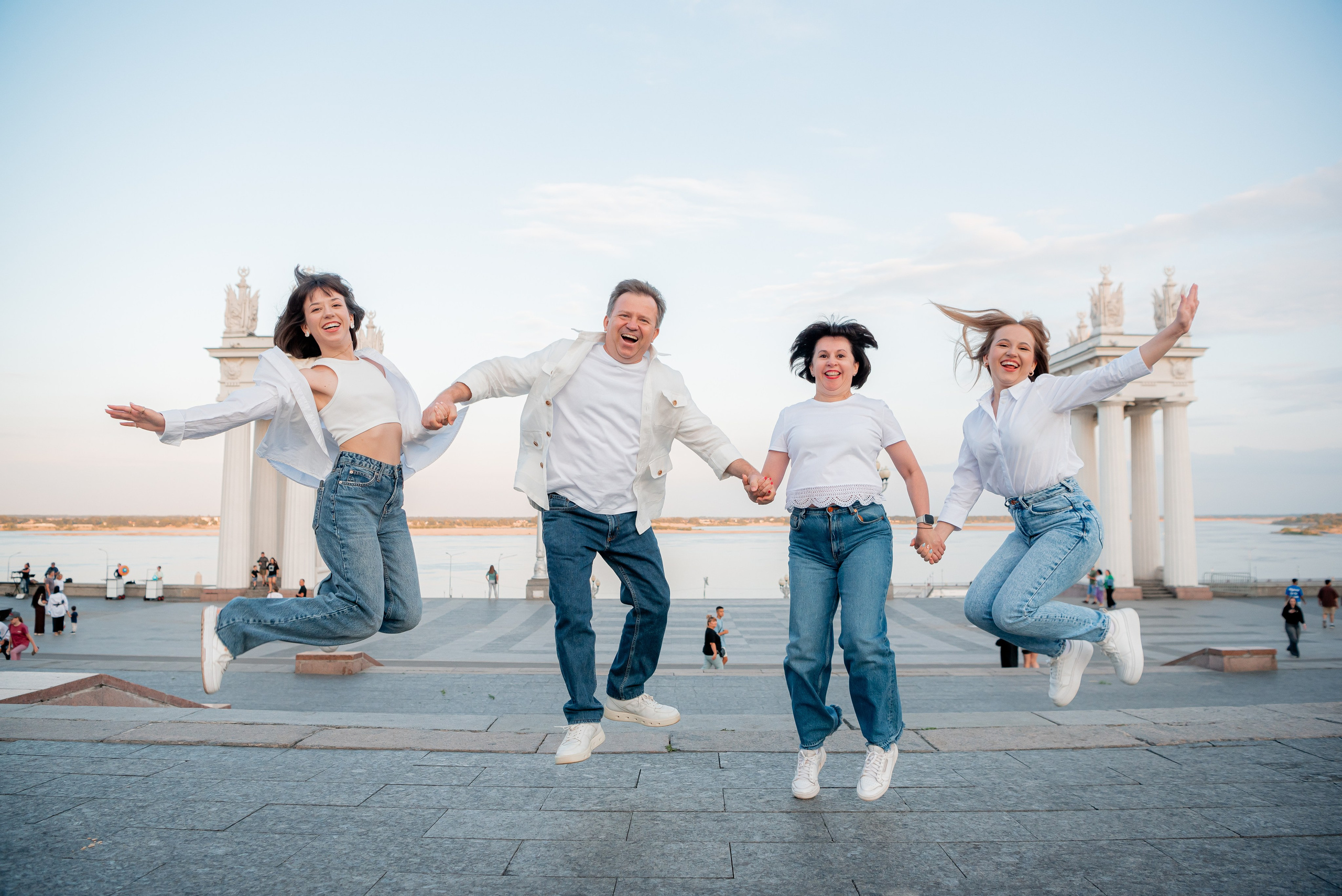Весёлые семейные фотографии где папа с мамой и две взрослых дочери подпрыгивают взявшись за руки на набережной Волгограда на фоне реки Волга