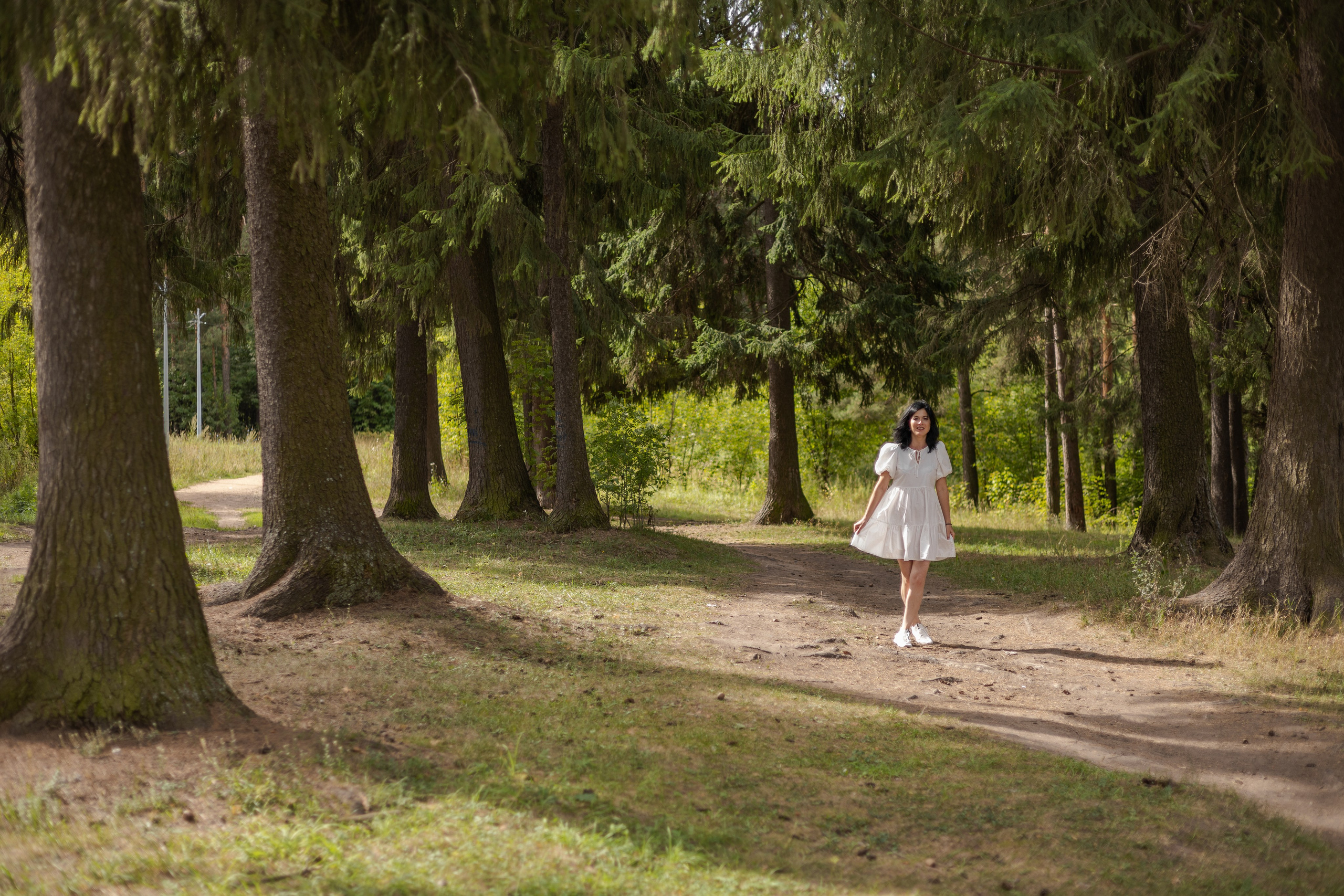 Девушка в белом платье в парке идет по тропе между сосен