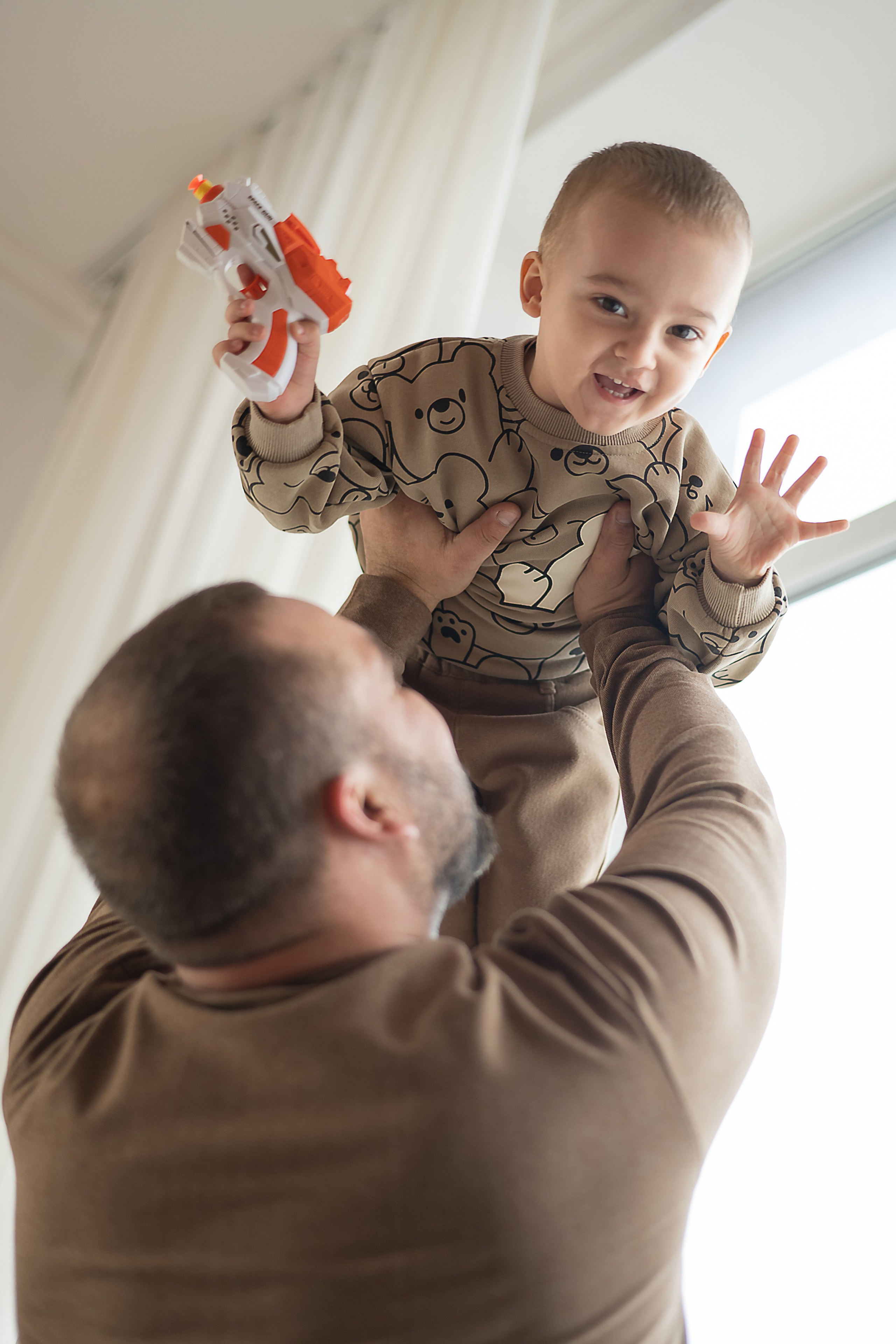 Папа подбрасывает счастливого малыша над головой на фоне окна