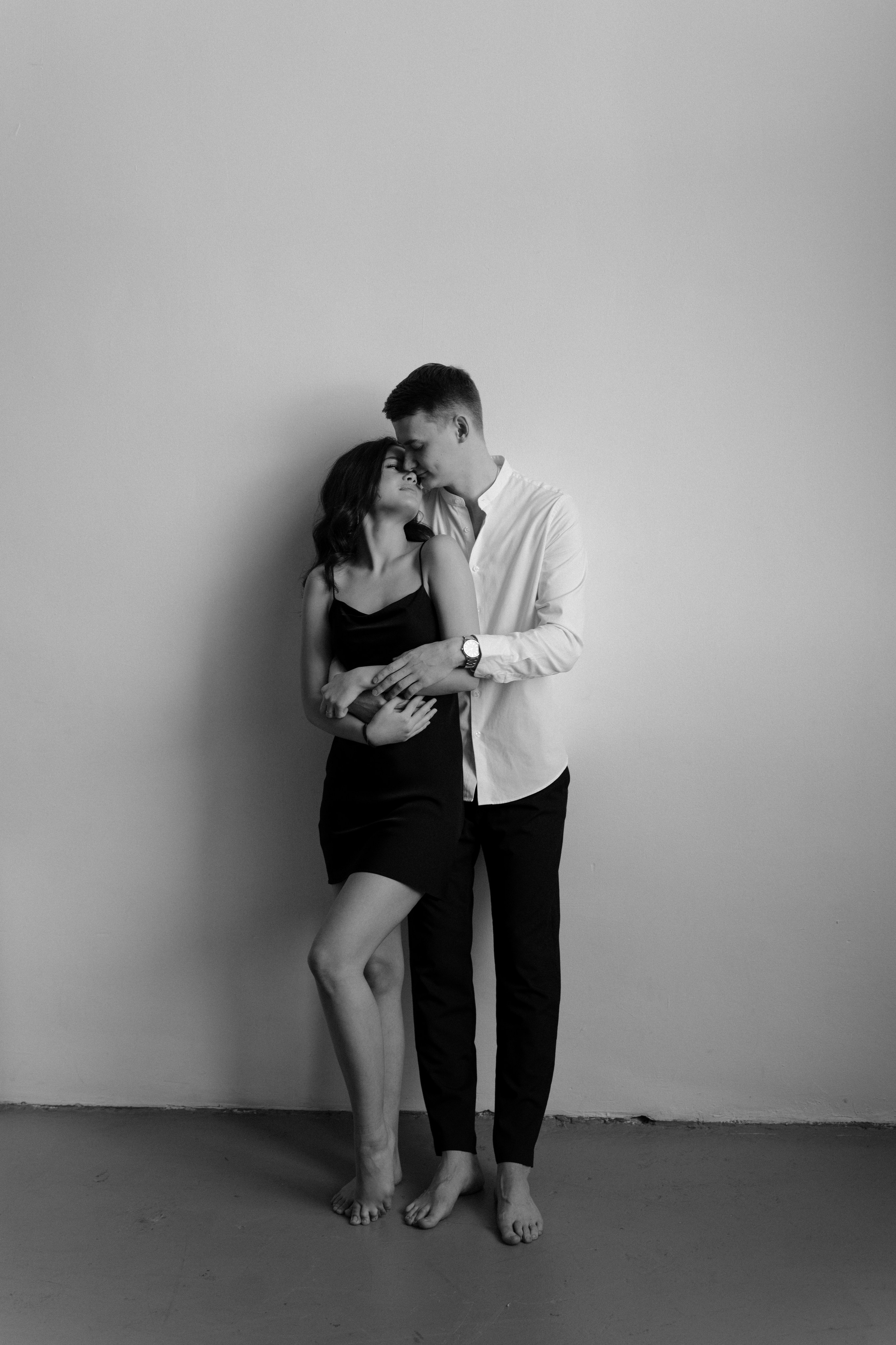 Девушка в коротком черном платье и парень в белой рубашке стоят у стены целуются в фотостудии.