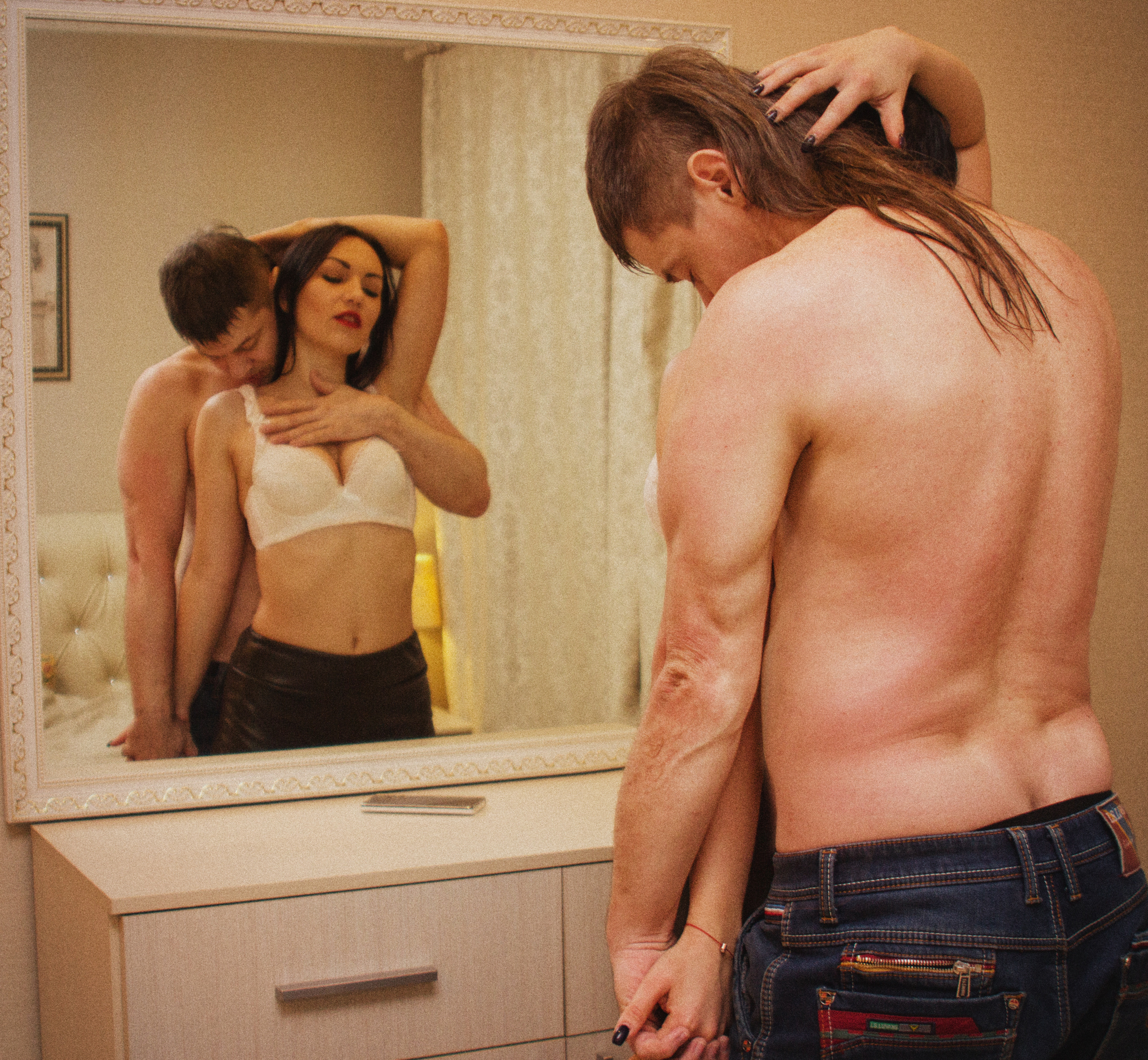 мужчина со  спины  обнимает женщину. женщина  смотрит на себя   в  зеркало. на ней белое  белье.