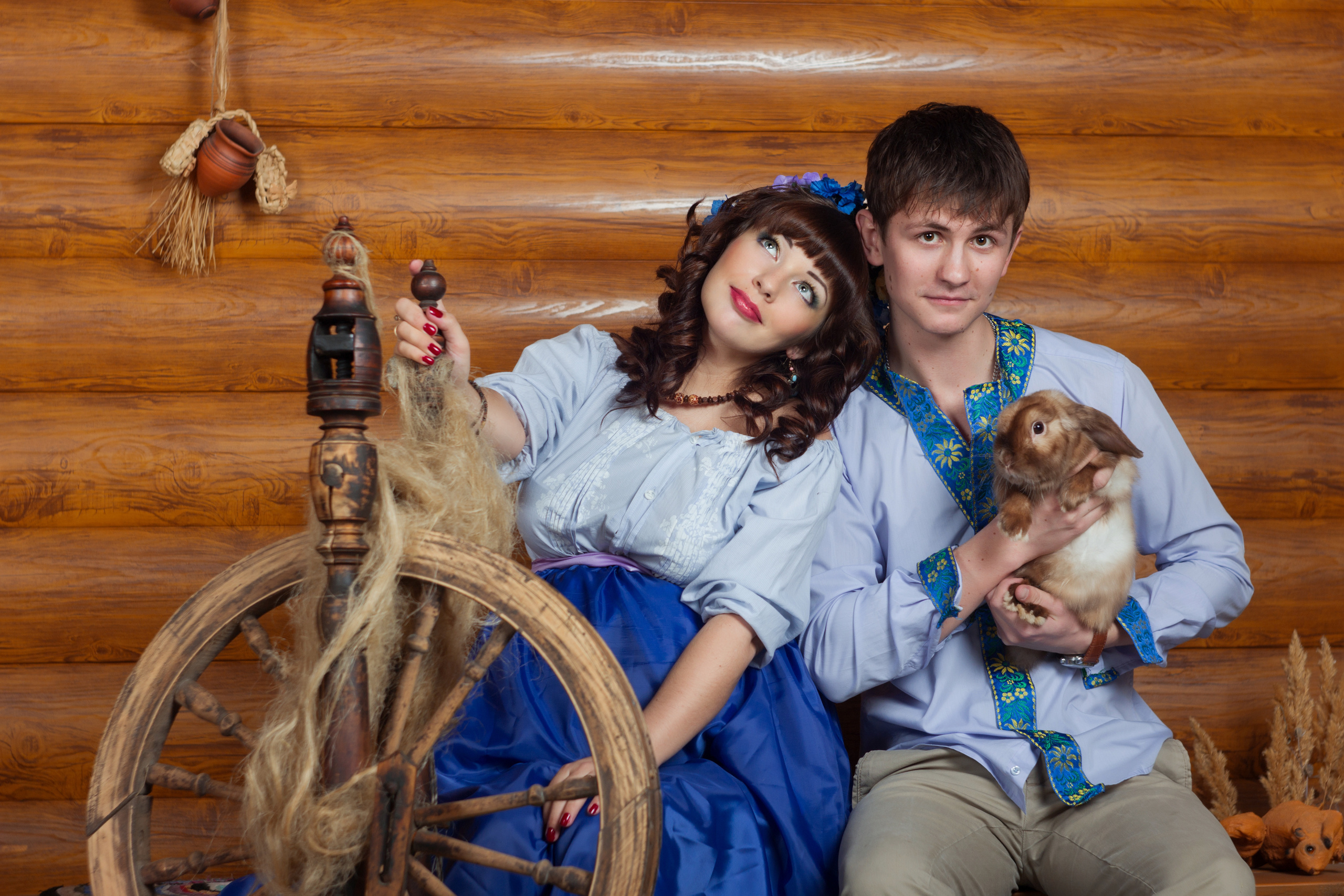 Парень  и девушка   в народных  костюмах сидят  возле  прялки. Парень держит  в руках кролика.