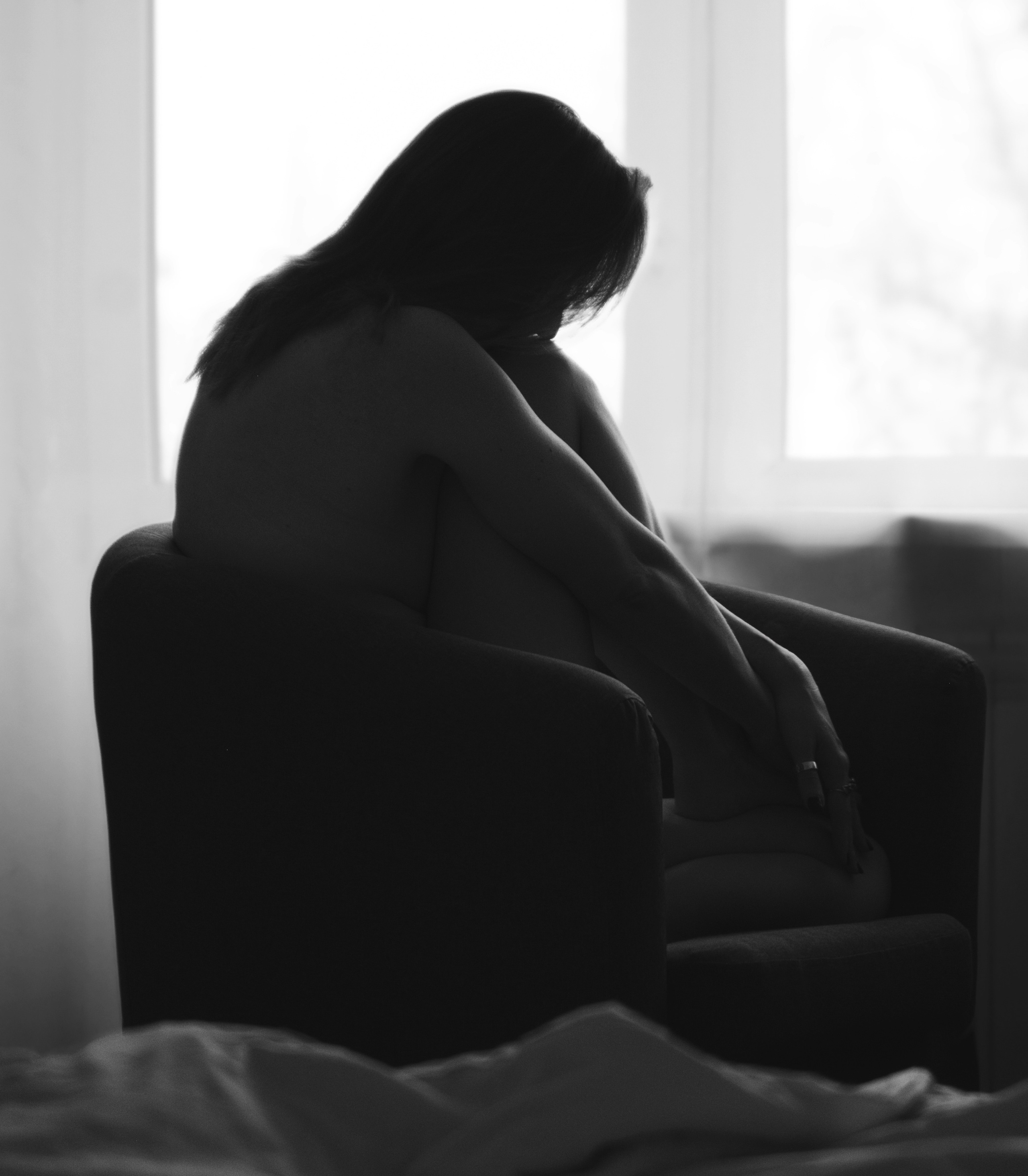 девушка сгруппировавшись сидит в кресле напротив яркого окна