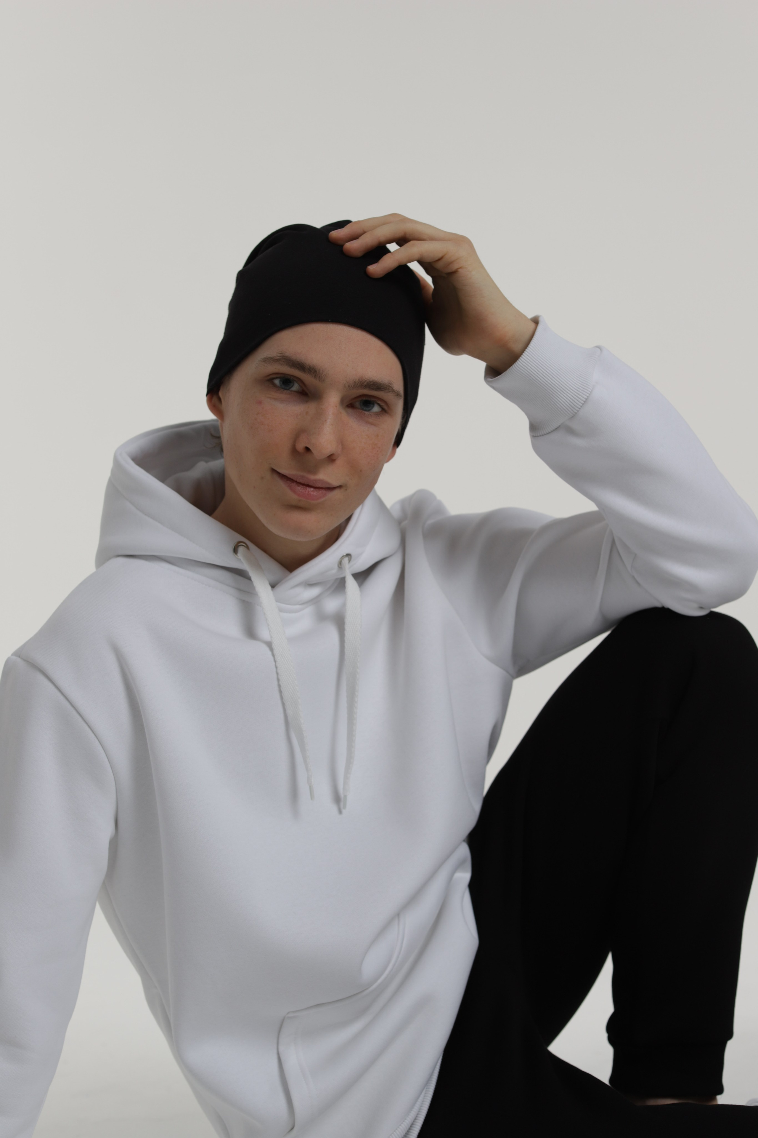 Молодой парень модель позирует в белом худи черной шапке на сером фоне
