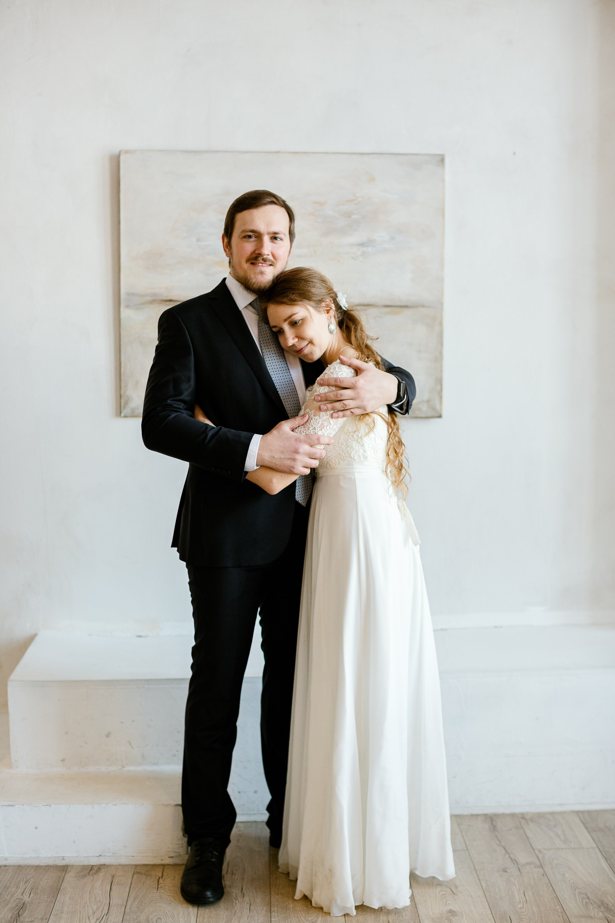 Жених и невеста в праздничной фотостудии. Жених в костюме, невеста в красивом белом платье. На фоне картина.