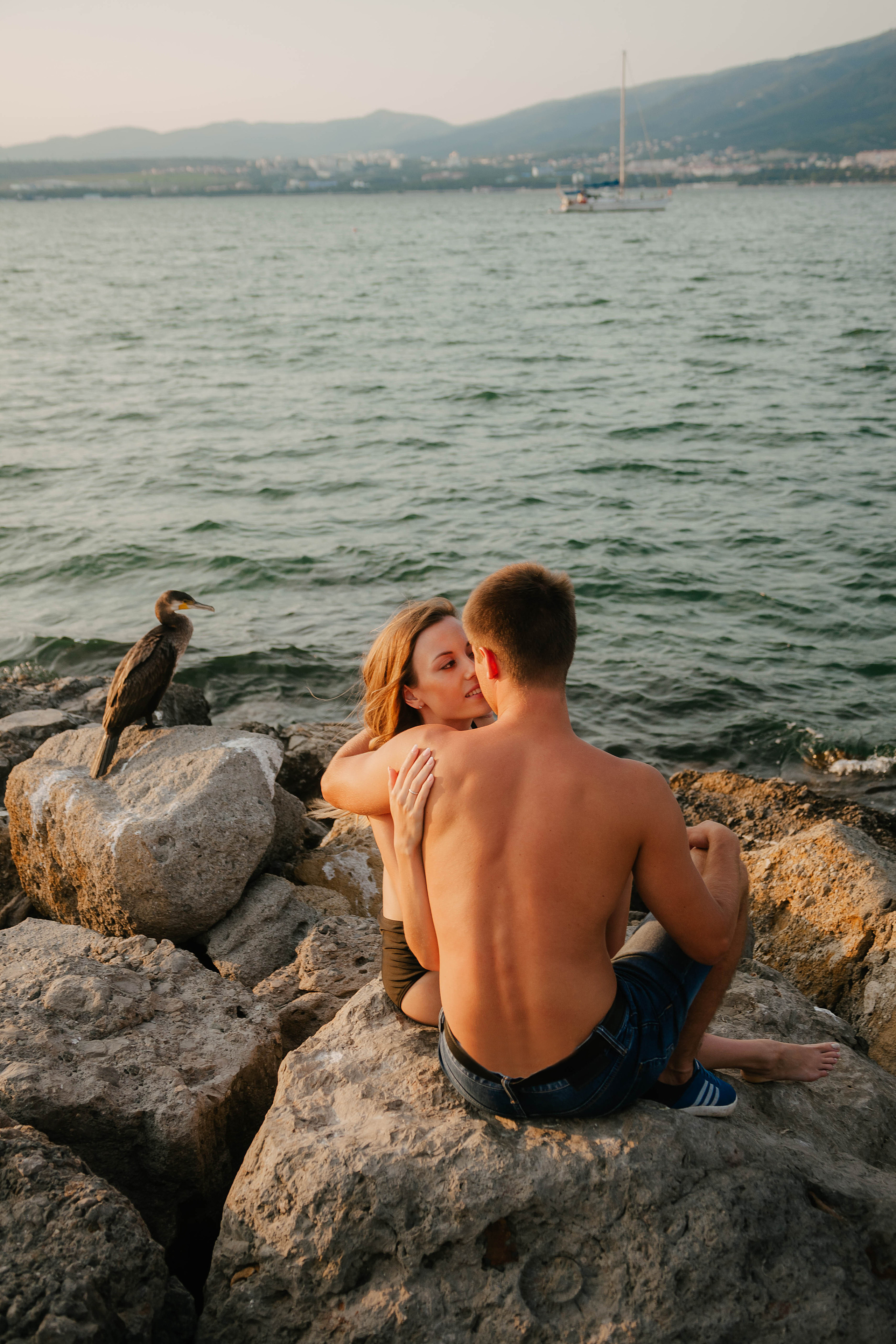 парень и девушка сидят на камнях на пляже рядом с ними сидит баклан