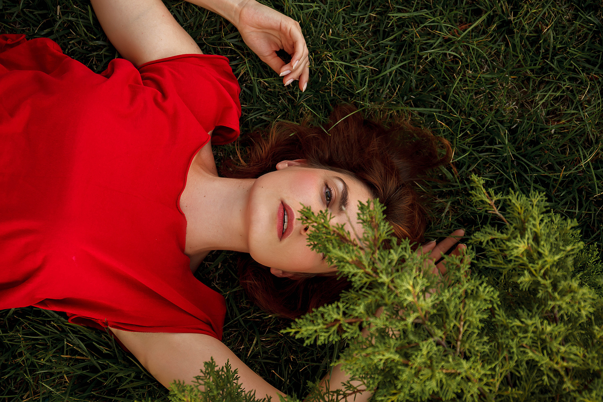 девушка в красном лежит на траве и прикрывает кустом один глаз