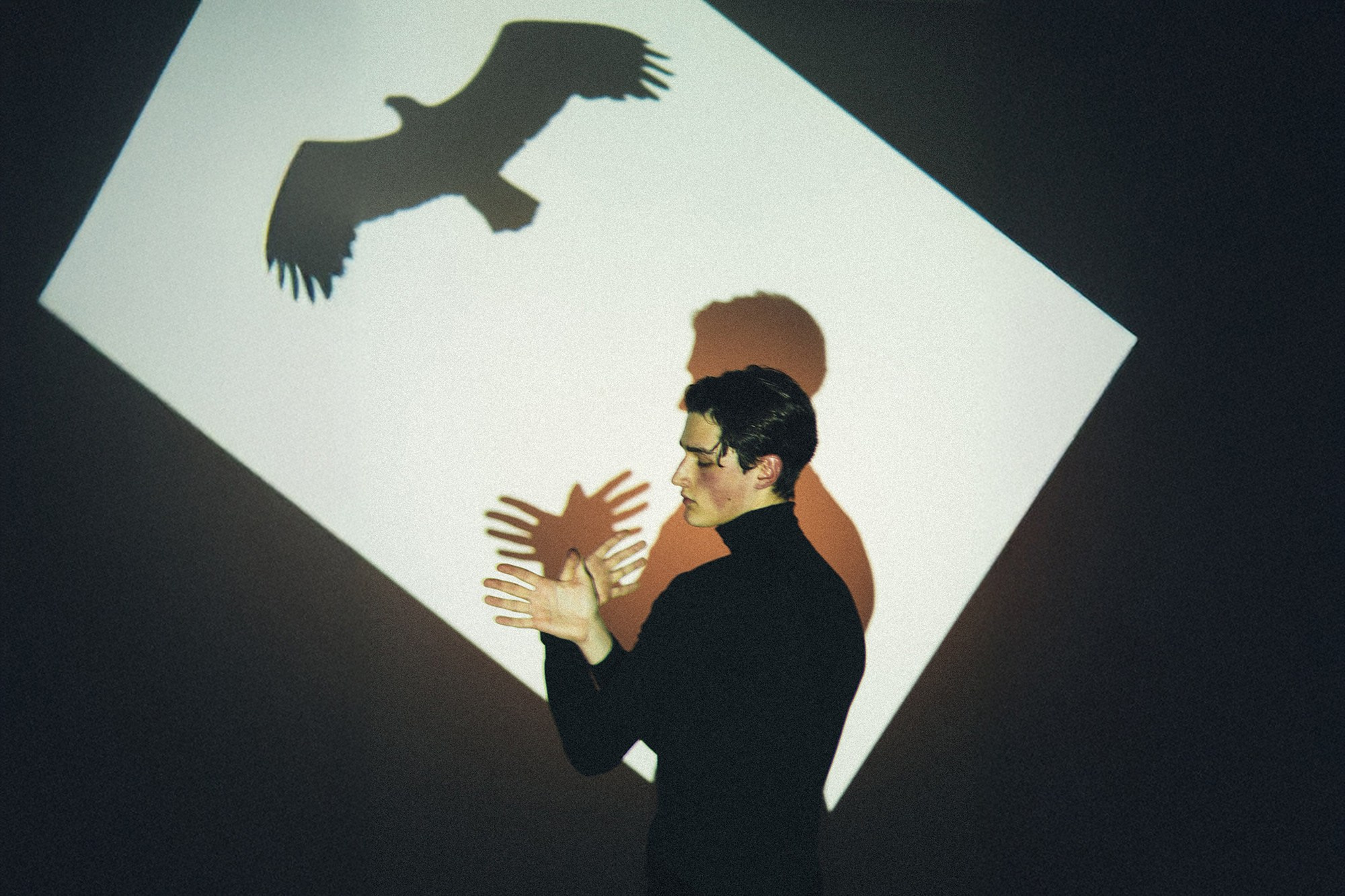 Творческий портрет от фотографа Сочи Галины Скорик. Молодой человек в черной водолазке на фоне светового пятна имитирует взмах крыльев, дополняя проекцию орла.