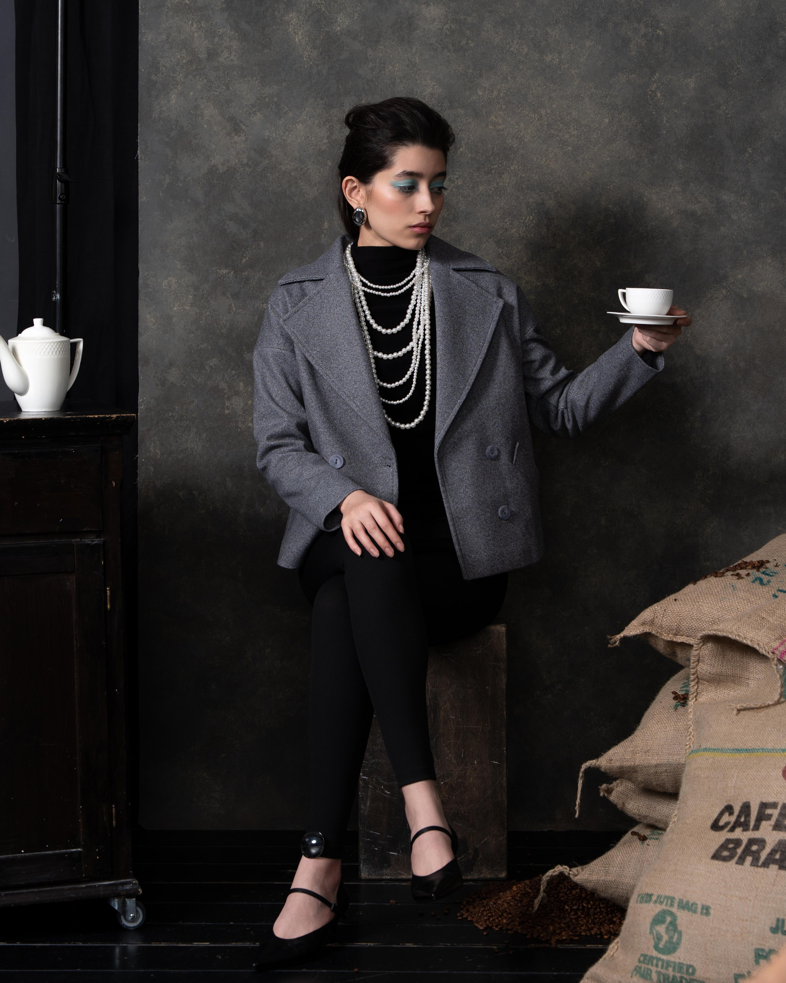 На изображении представлена женщина, сидящая на деревянном ящике в интерьере, который напоминает кофейню или студию. Она одета в серое пальто, черную водолазку и черные брюки. На шее у нее несколько нитей жемчуга. В руке она держит чашку с кофе. На заднем плане видны мешки с кофе и кофейный столик с чайником.