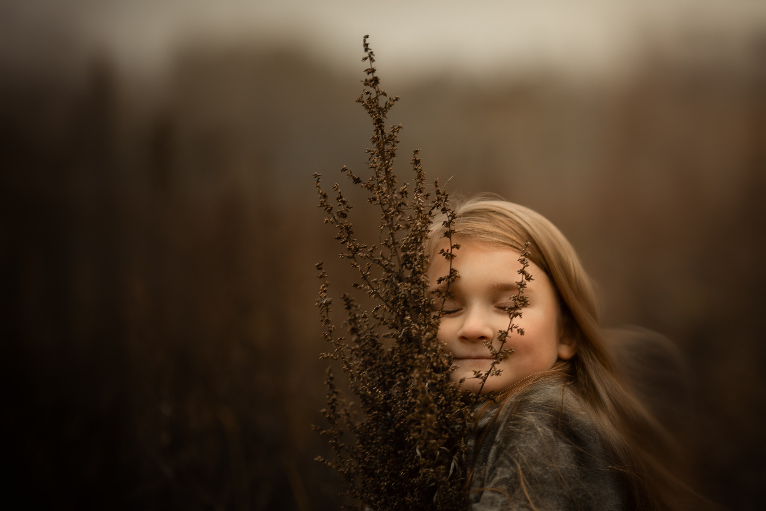 Девочка с закрытыми глазами обнимает сухое растение на фоне тёплого осеннего поля, мягкий свет и спокойная атмосфера.