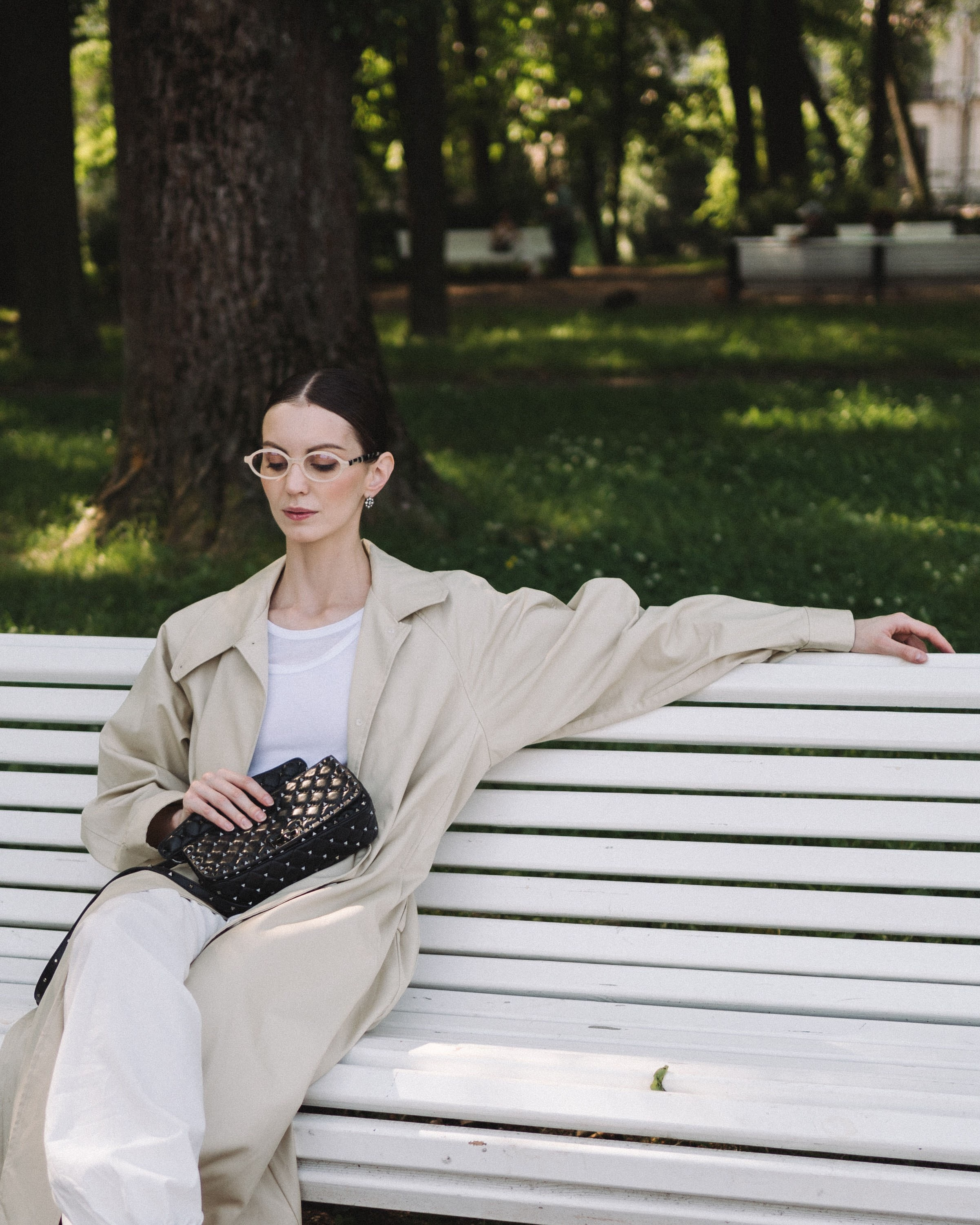 Атмосферная фотография, на которой модель сидит на лавочке в парке в модной одежде