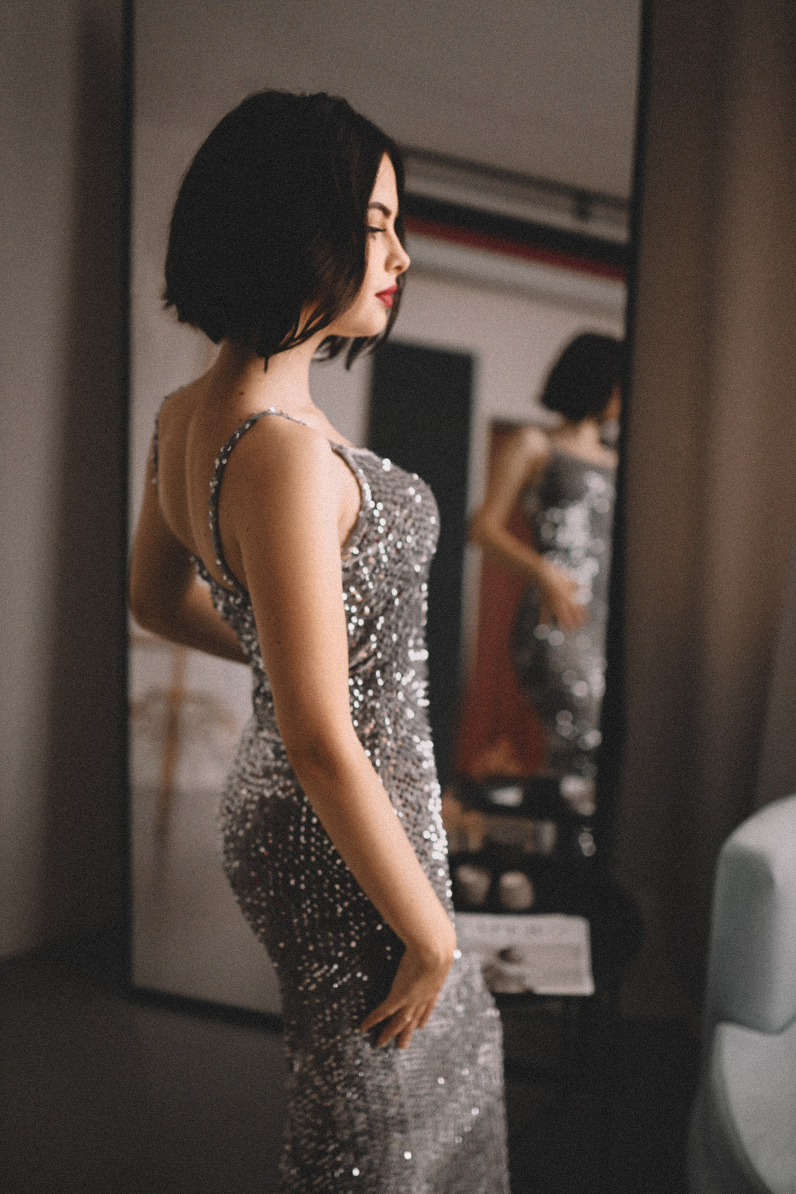 Девушка в блестящем платье стоит у зеркала, отражение усиливает глубину и символизм кадра.