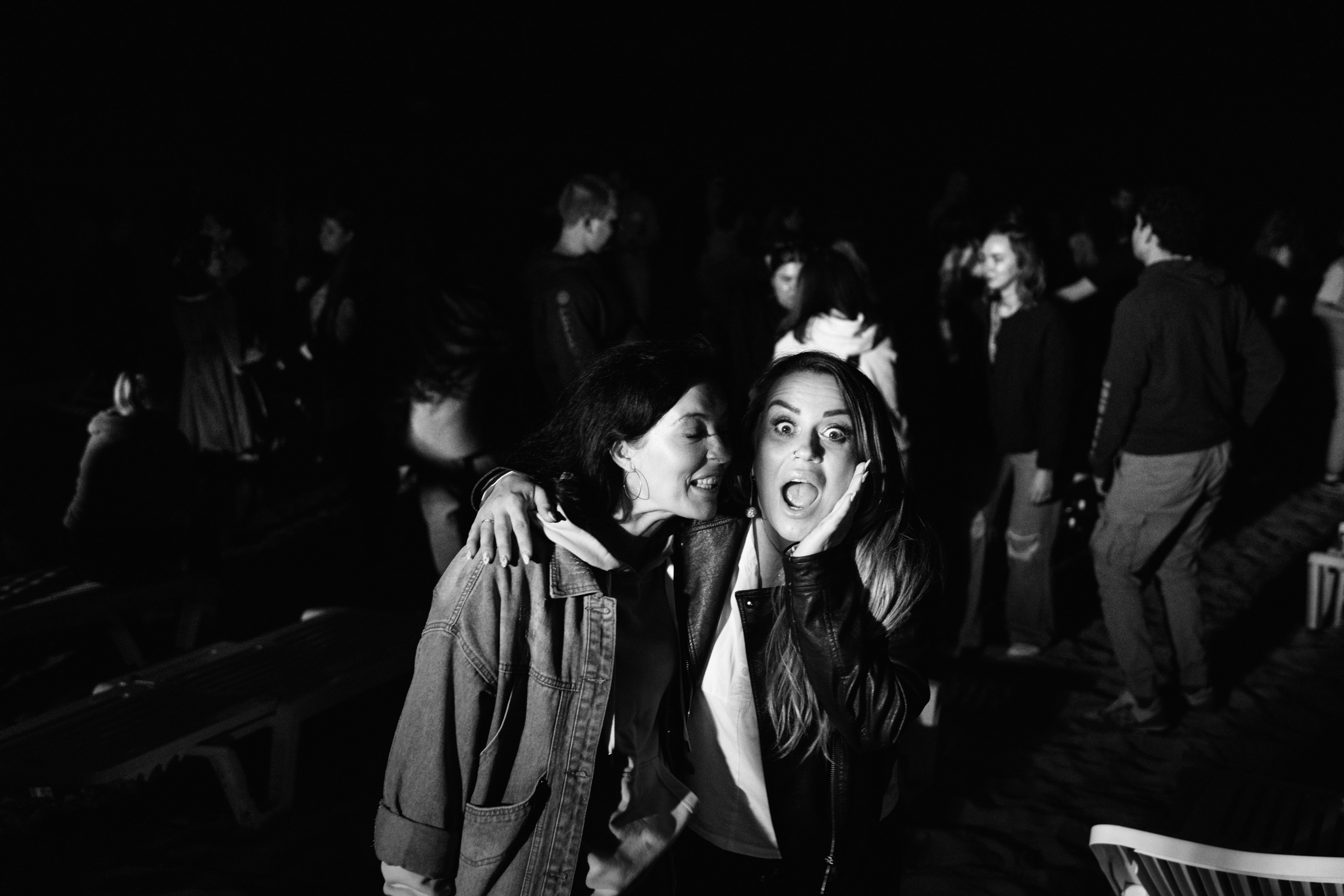 Две девушки позируют вместе, одна смотрит в камеру c открытым ртом, вокруг — люди ночью (ч/б фото)