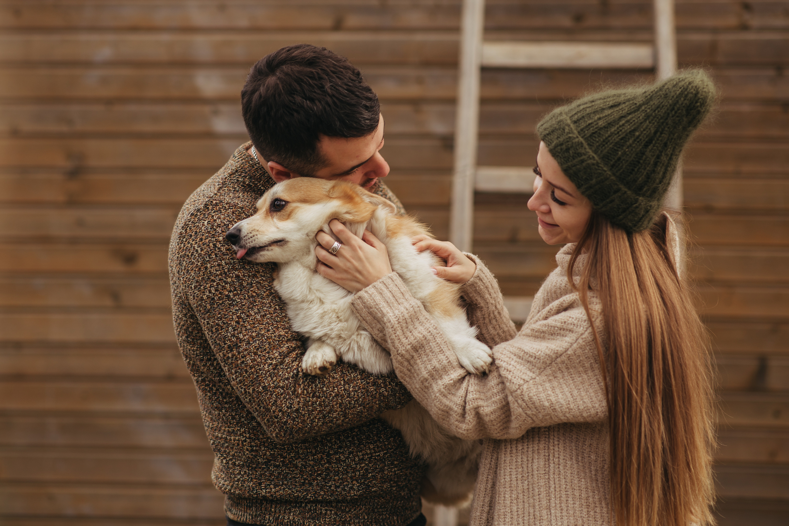 Девушка с длинными волосами в бежевом свитере и в зеленой шапке гладит собаку, которая сидит на руках у мужчины в коричневом свитере. На заднем плане лестница