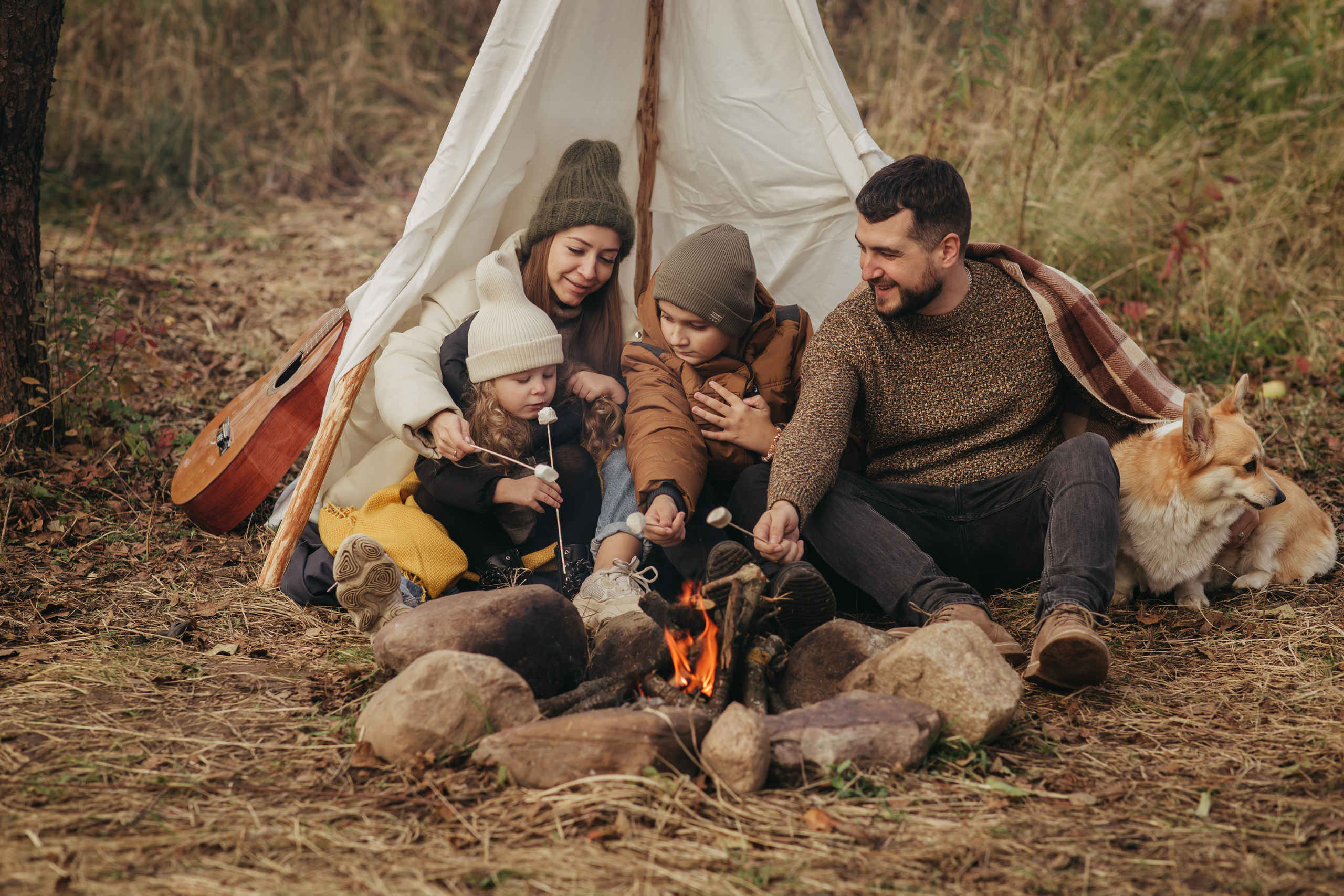 Семья в бежевой одежде сидит у костра и жарит маршмелоу. Шалаш