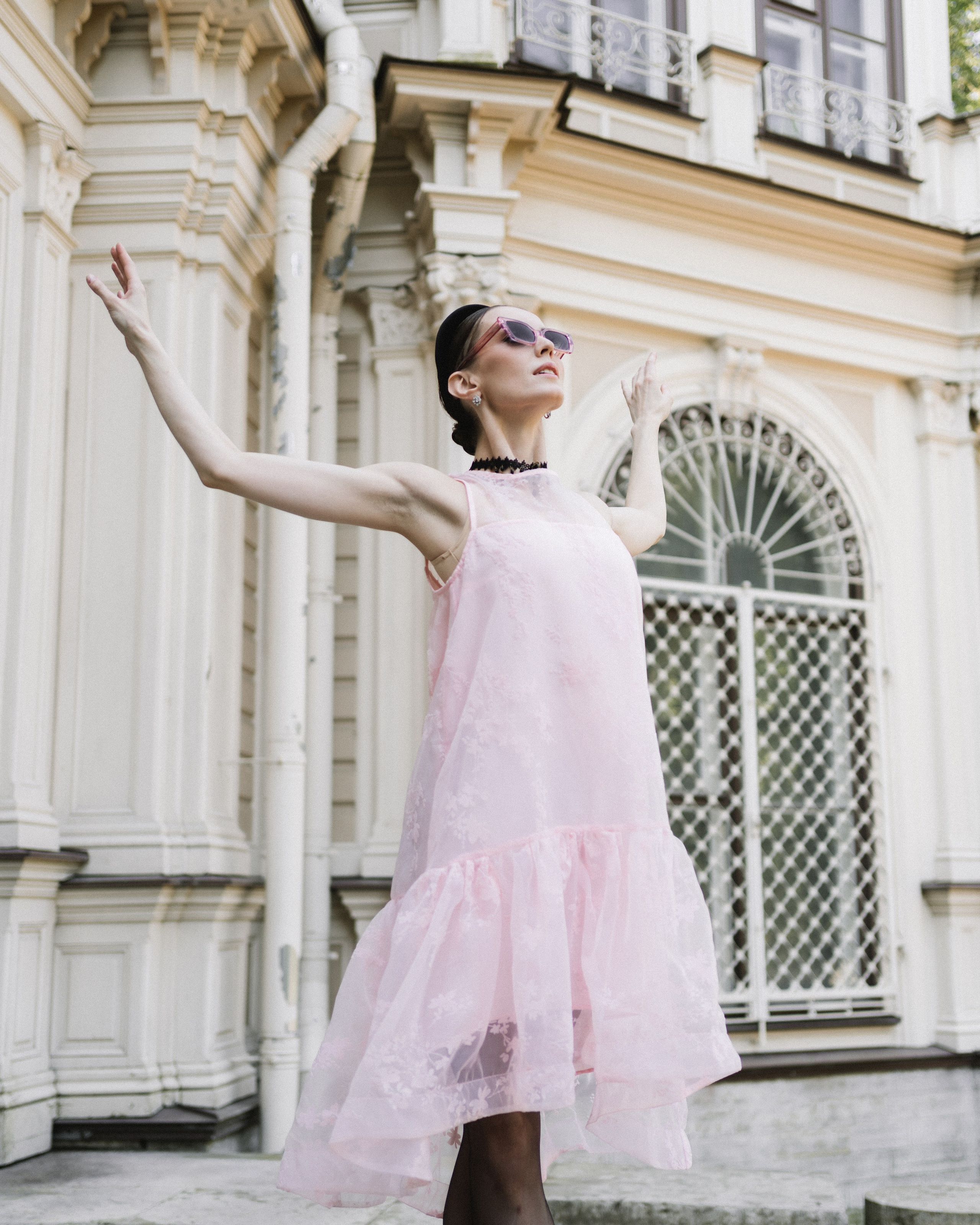 Динамичная фотография, на которой модель изображает балерину. Она стоит на фоне красивой архитектуры. Одета в розовое воздушное платье, очки. 