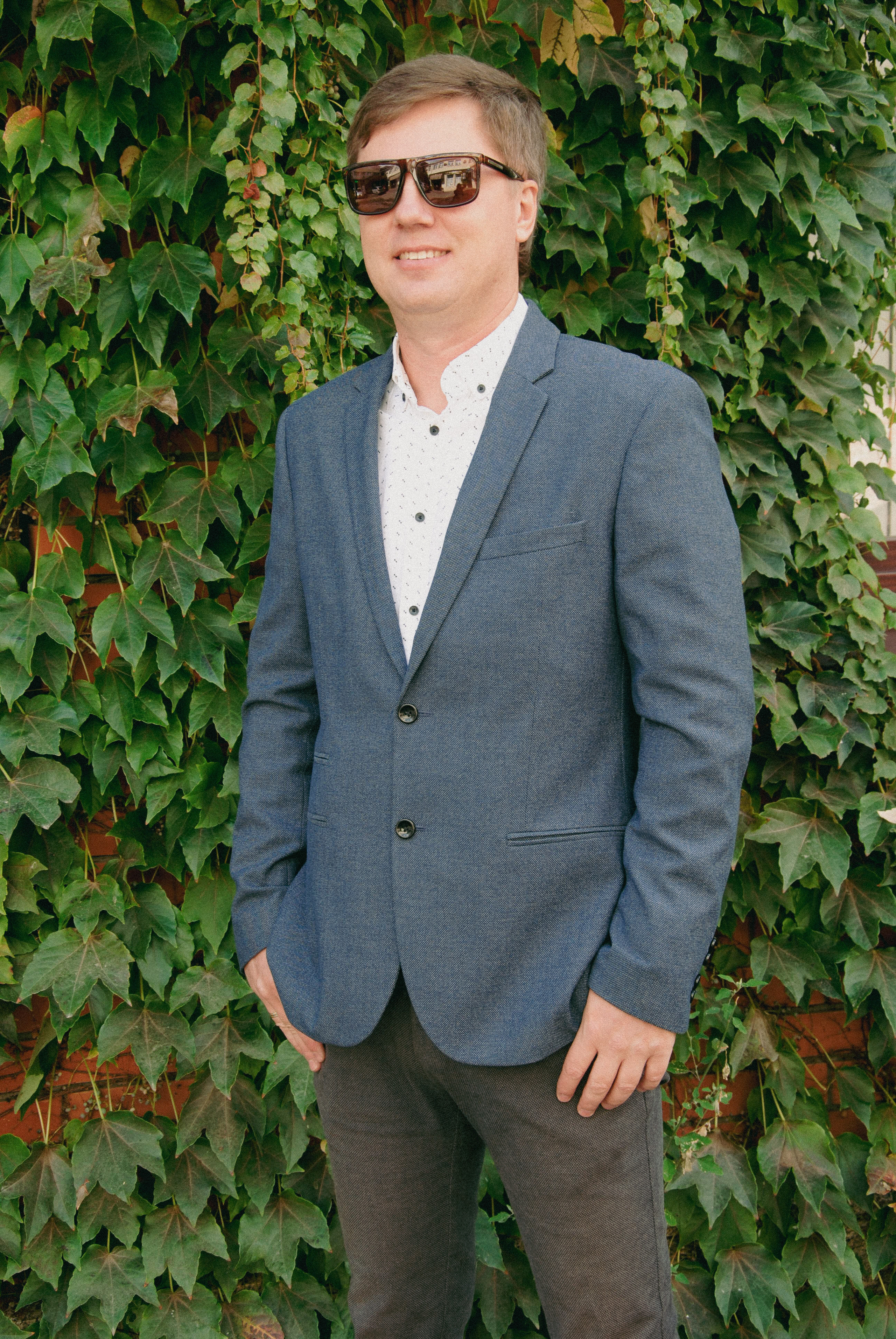 Молодой человек в сером пиджаке стоит на фоне зеленого плюща