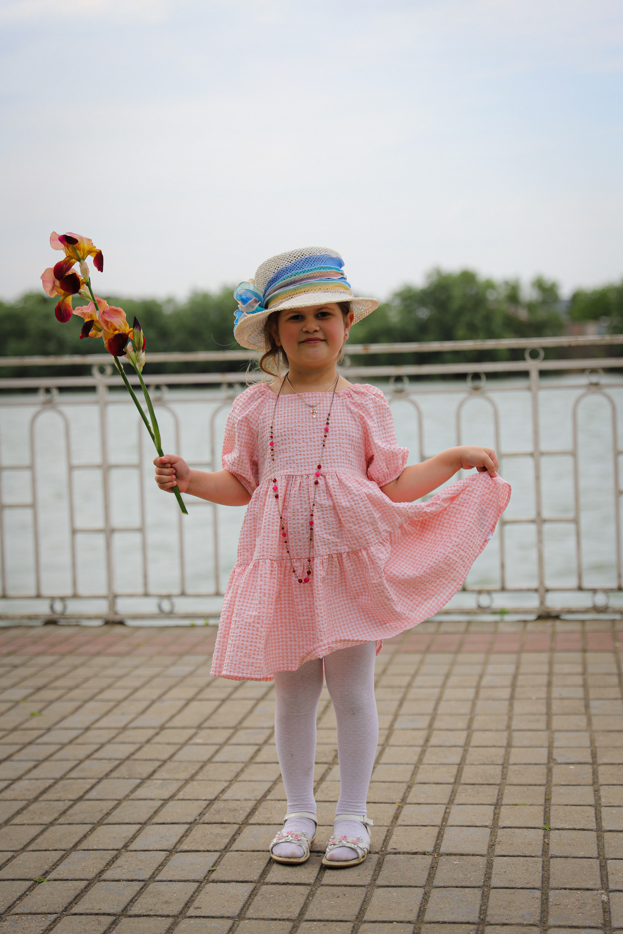 Веселая девочка в шляпе с цветком позирует в розовом платье на фоне реки Кубань на набережной